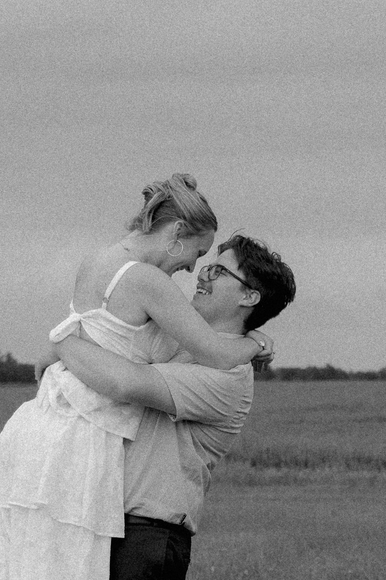 Black and white film photo of joyful couple embracing in open field, man lifting smiling woman.