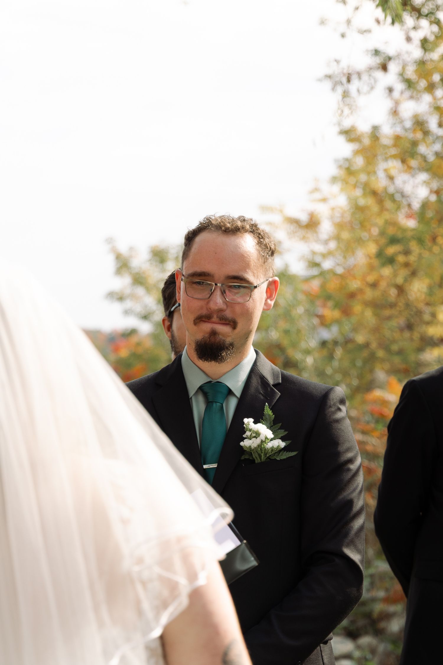 Emotional groom in black suit with teal tie and boutonniere watching bride approach during outdoor ceremony.