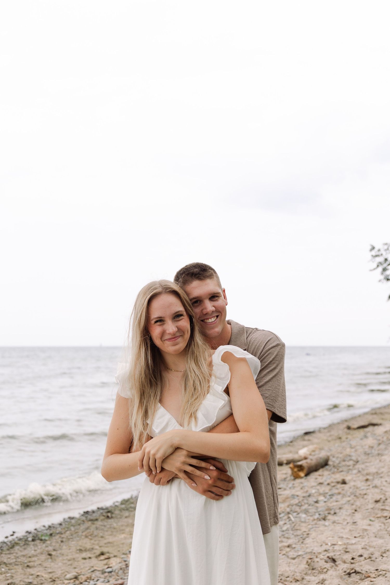 Smiling couple embracing on a pebble beach with calm ocean water in the background.