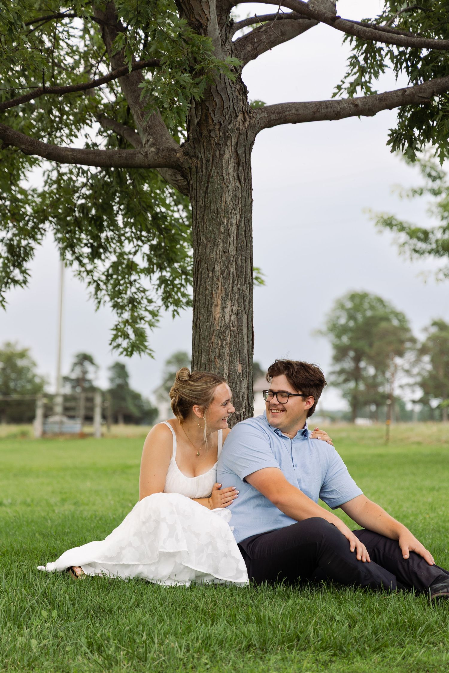 Engaged couple sitting together on lush green grass beneath a large tree, smiling at each other.