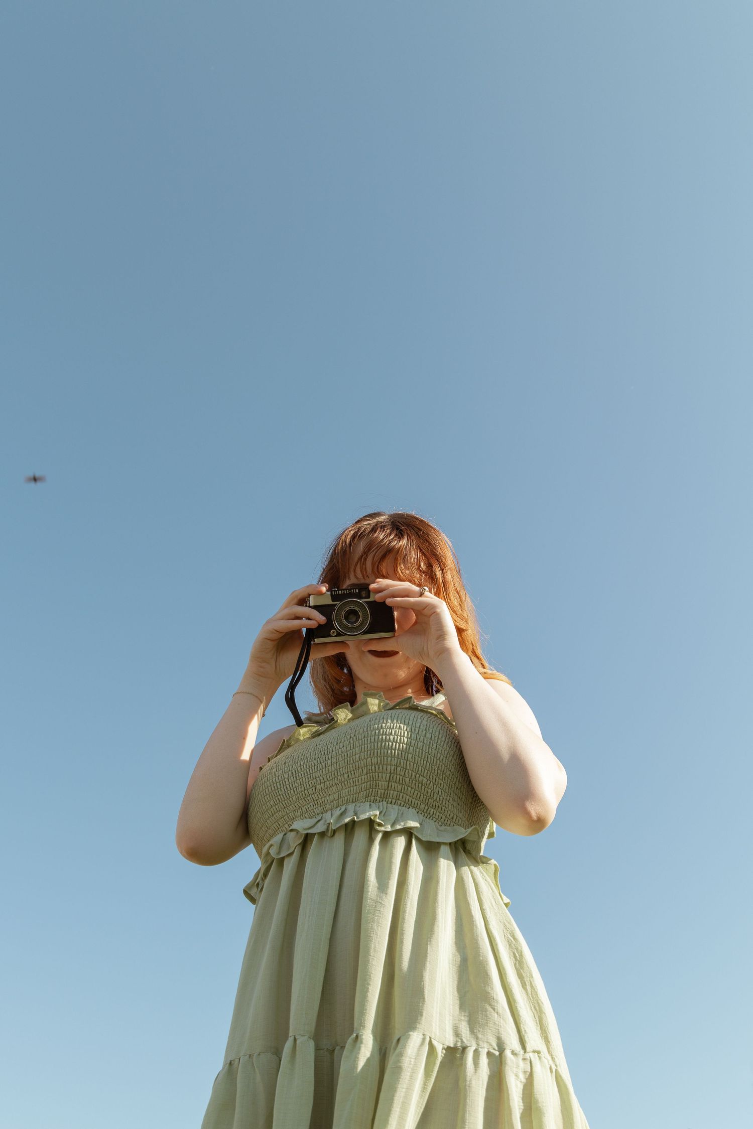 Young woman in green dress photographing with film camera against clear blue sky outdoors.