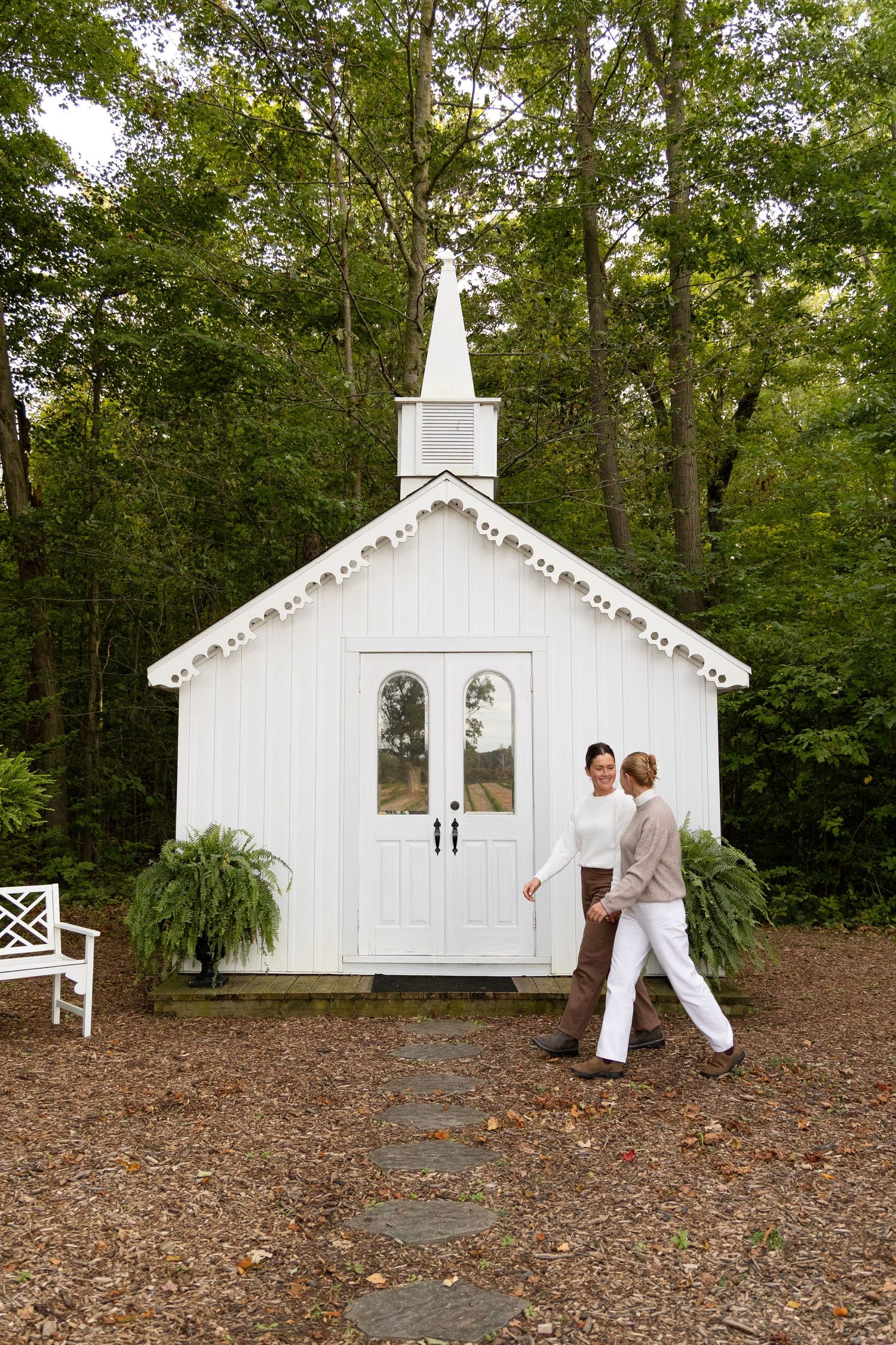 Two women holding hands walking toward a charming small white chapel surrounded by lush green trees.