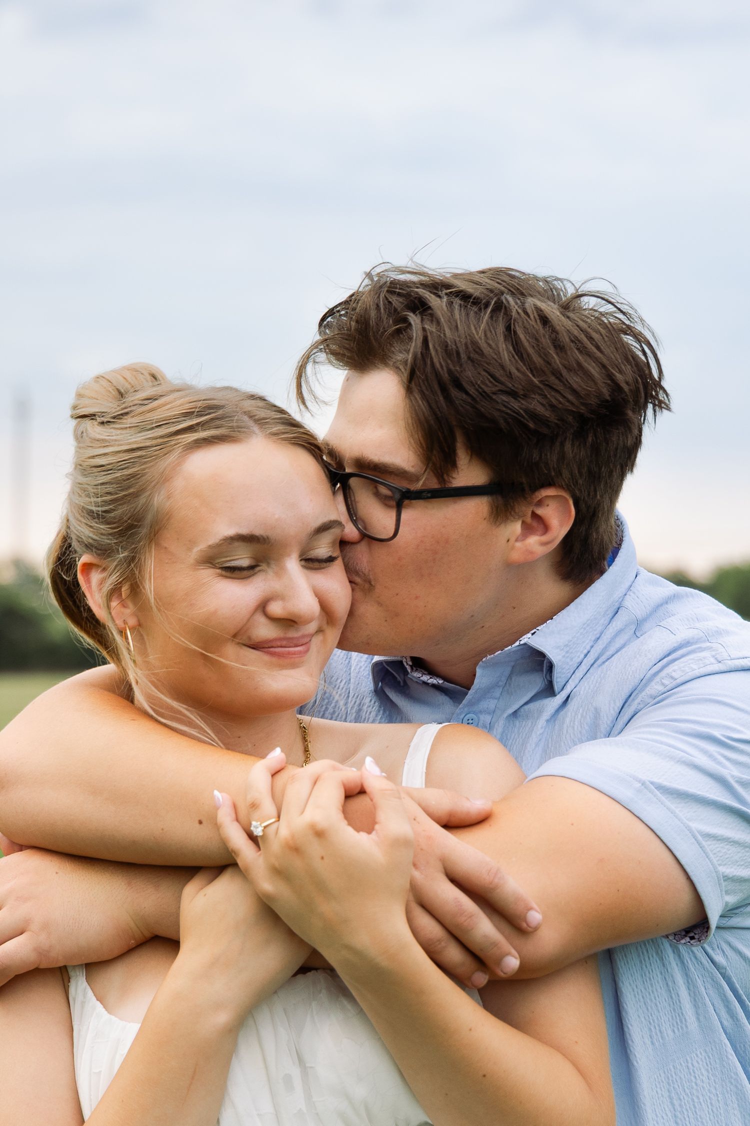 Young couple sharing a tender kiss, man in blue shirt embracing woman from behind showing engagement ring.
