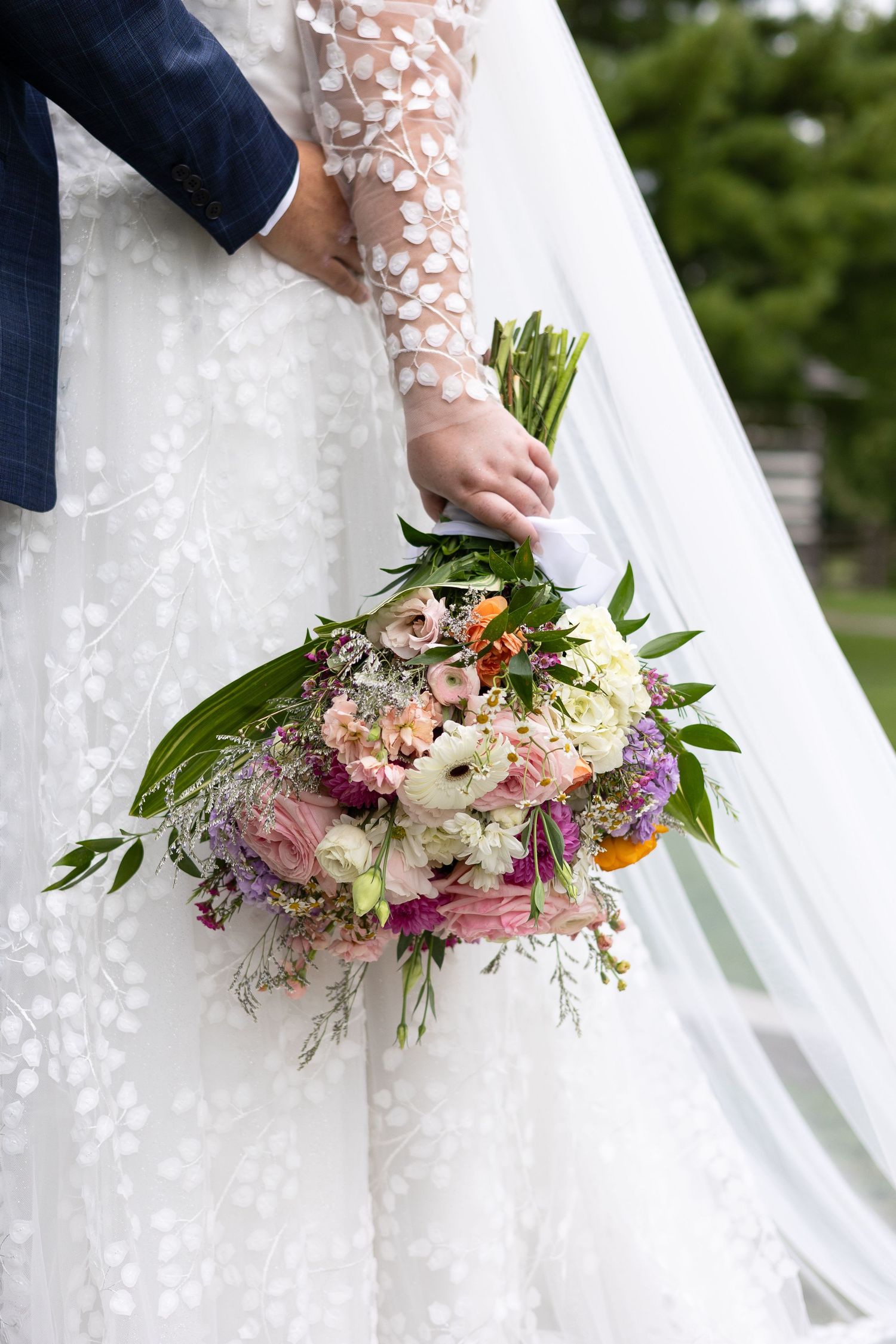 Bride in lace gown holds colorful wedding bouquet of pink roses and wildflowers beside groom.