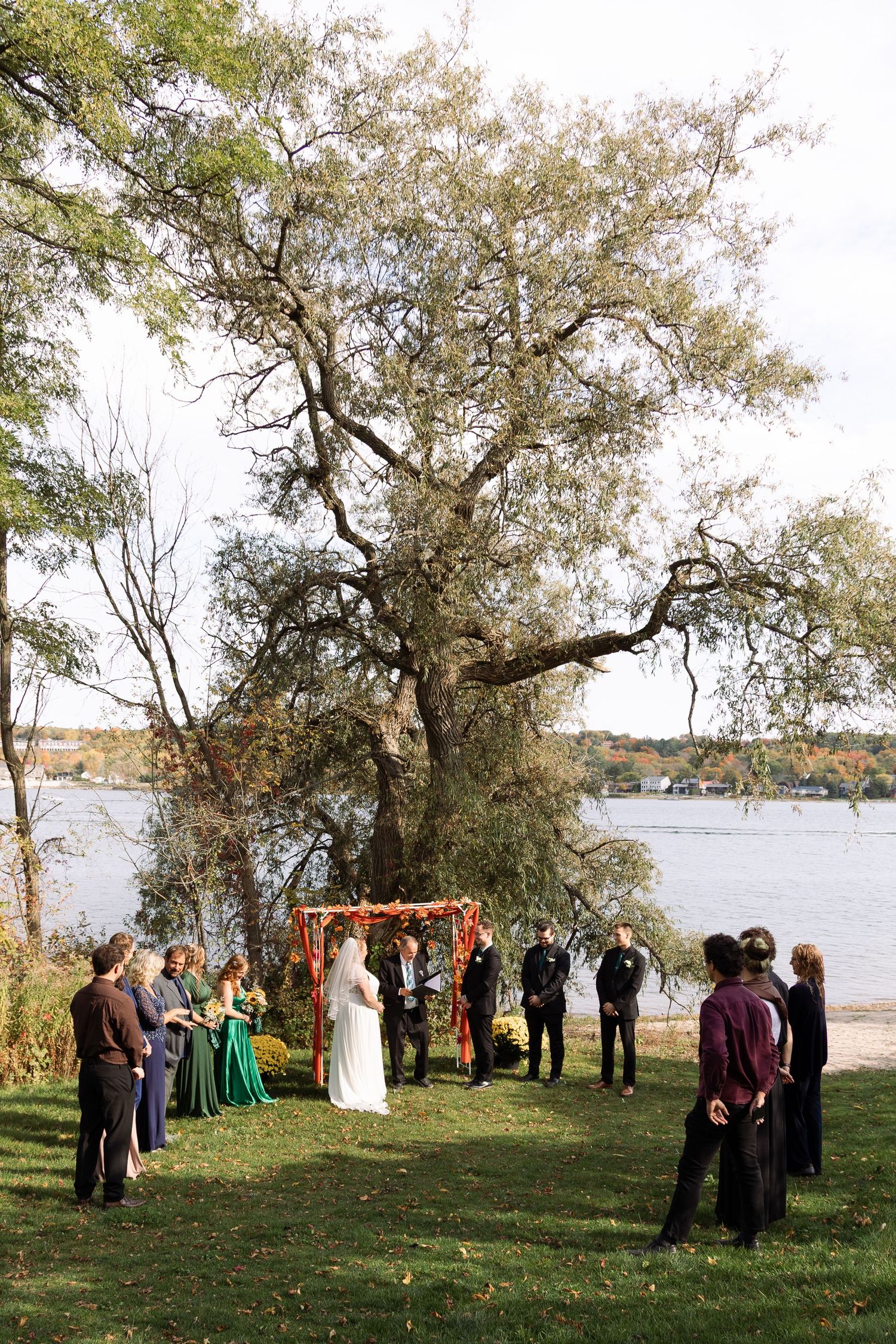Small intimate outdoor wedding ceremony beside a lake in autumn with colorful fall foliage and a floral arch.