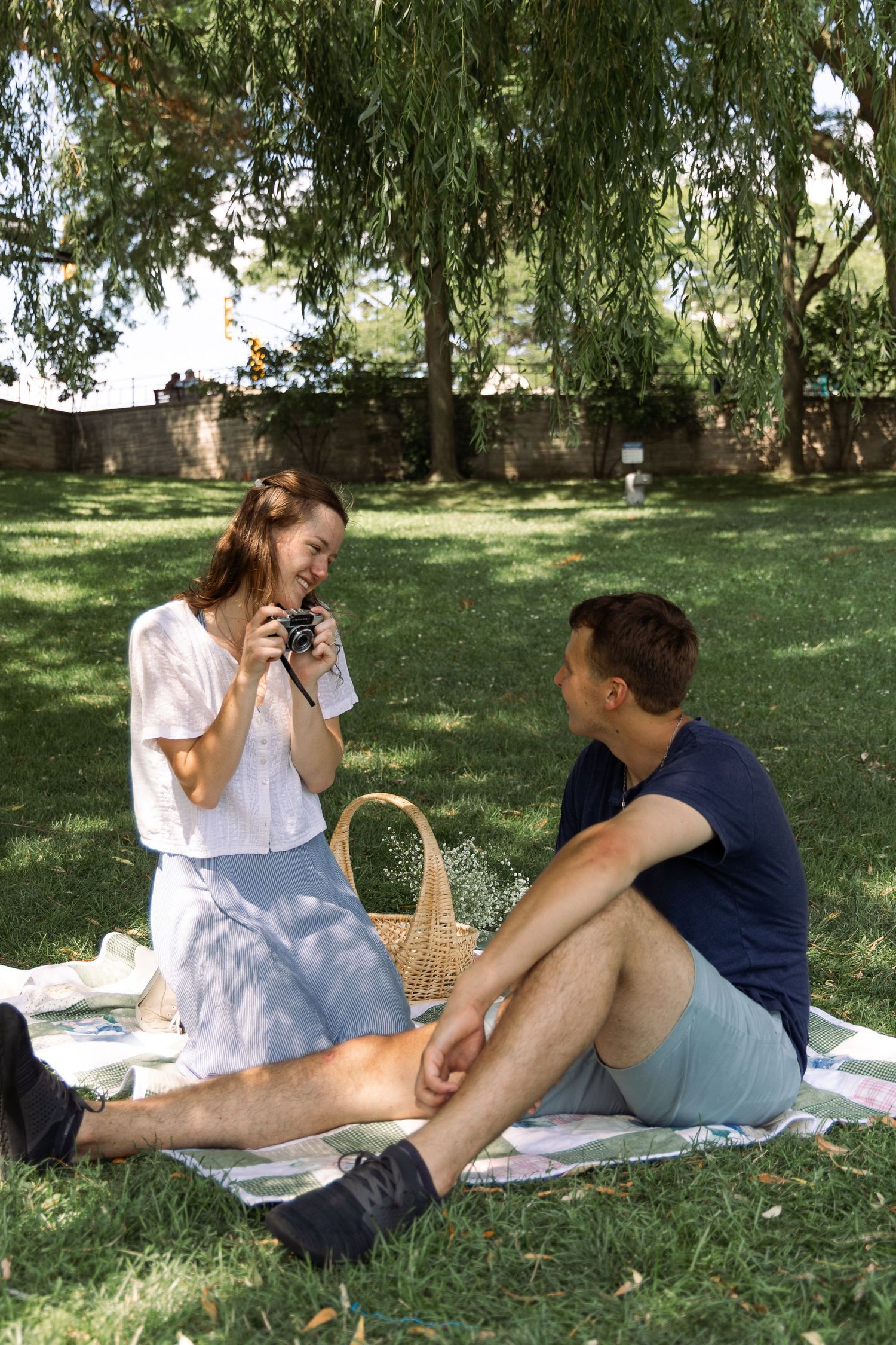 Young couple enjoying a sunny picnic on a blanket under willow trees in a park.