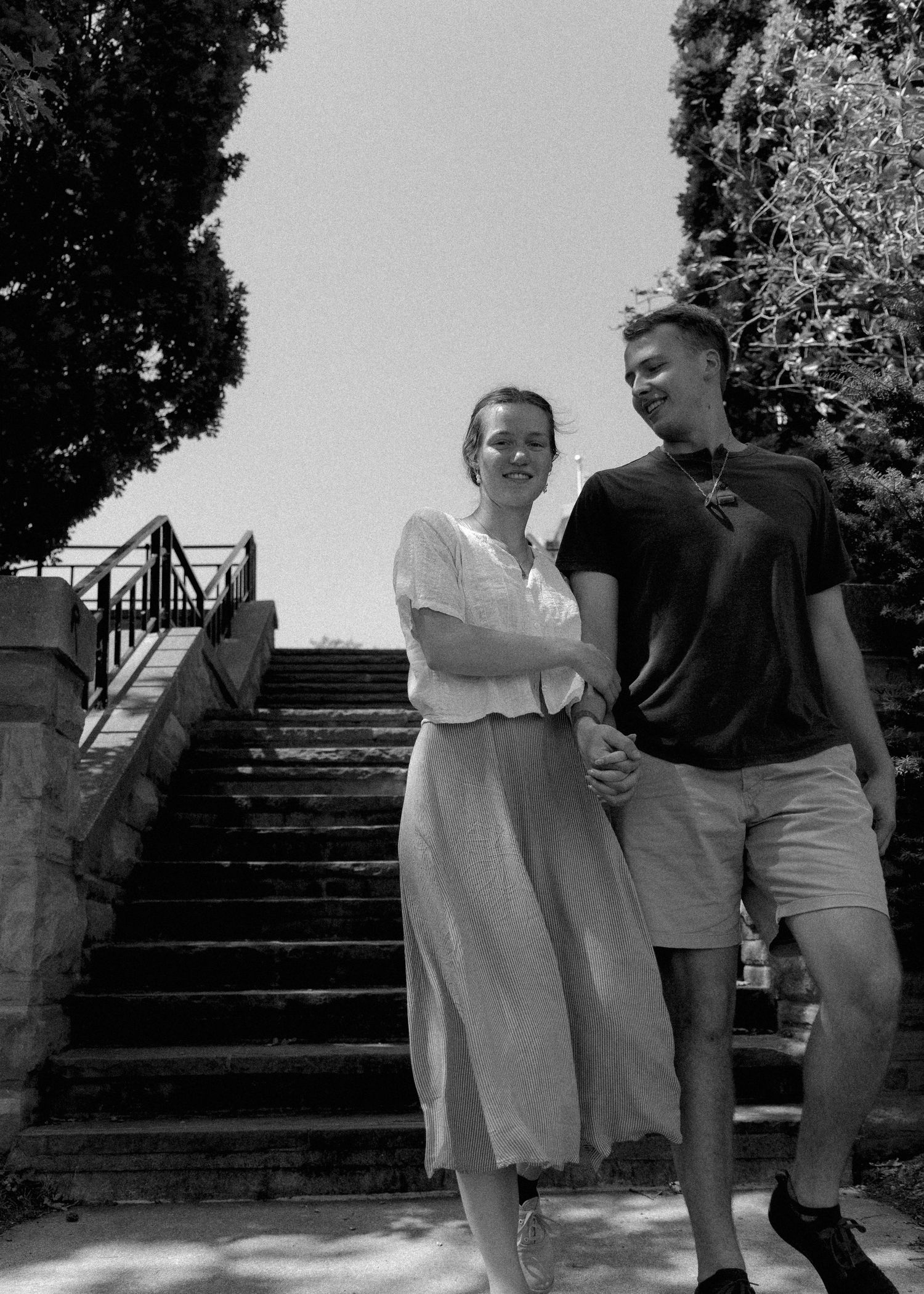 Smiling couple walks down outdoor stairway flanked by trees in black and white engagement photo.