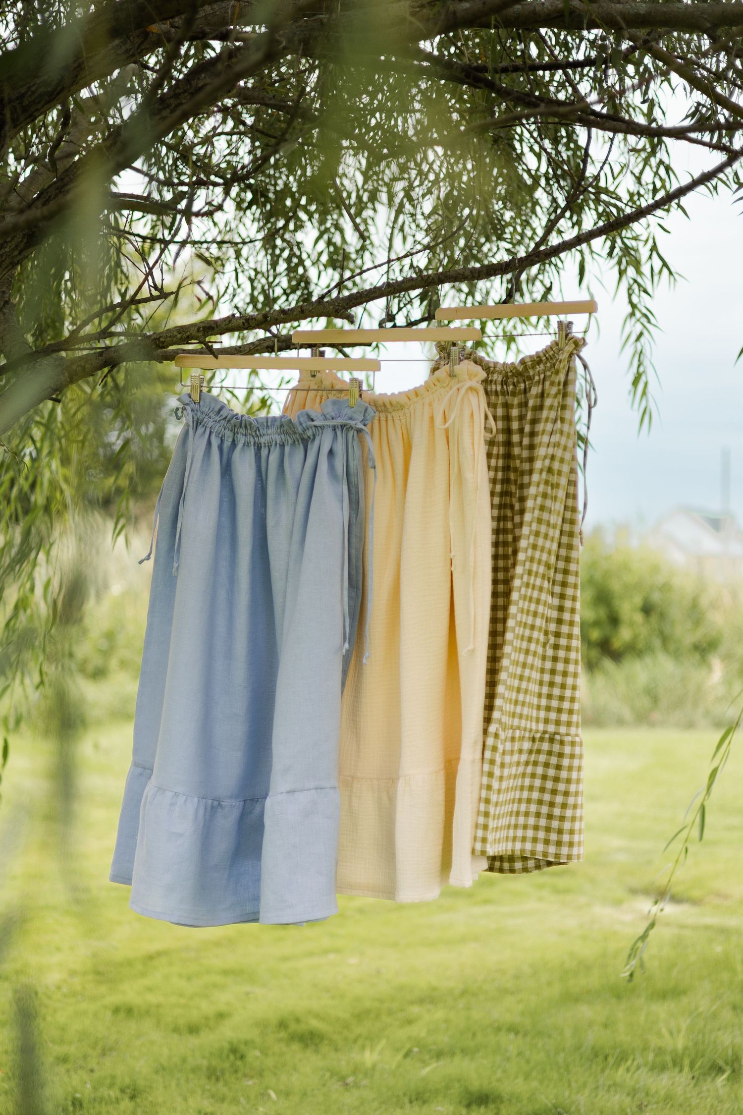 Three pastel midi skirts in blue, yellow, and green gingham hang on wooden hangers from a willow tree branch.