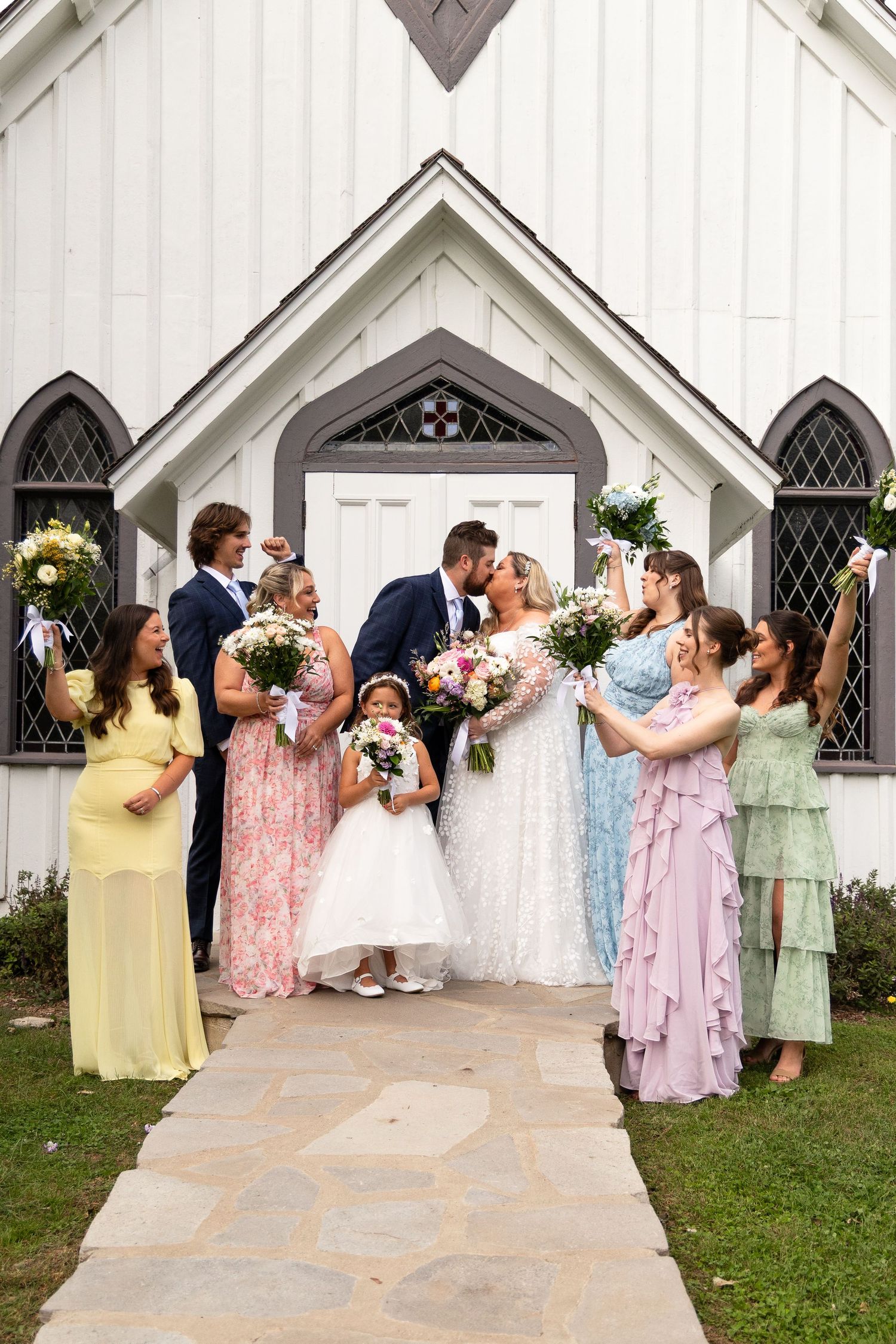 Wedding party celebrates outside white chapel as bride and groom share a romantic kiss.