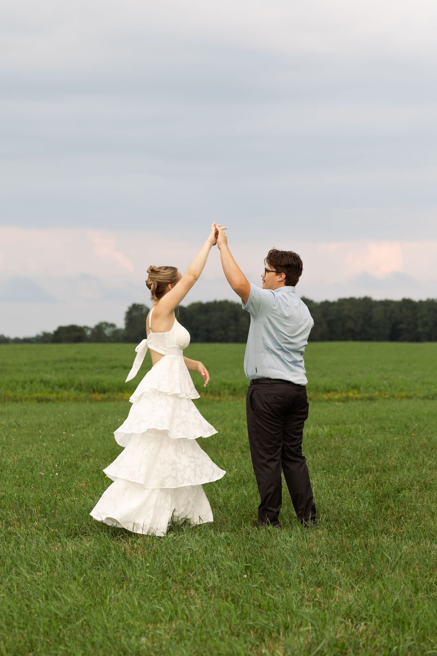 Bride and groom dancing in green meadow under cloudy sky, raising joined hands together outdoors.