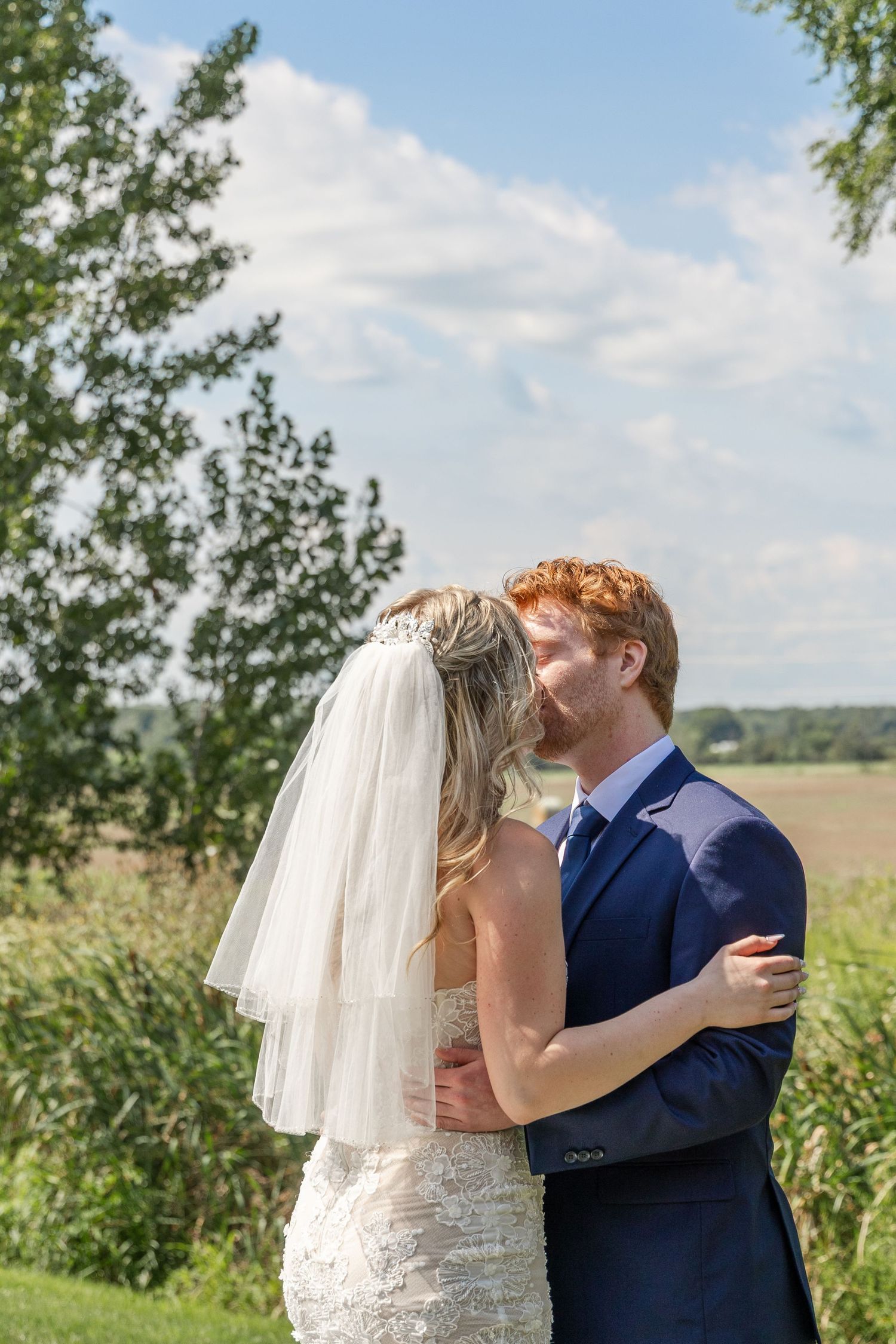 A groom in a navy suit kisses his bride wearing a lace dress and veil outdoors on a sunny day.
