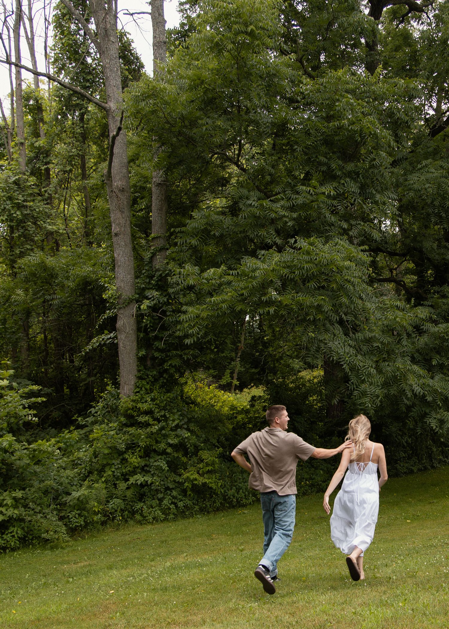 Couple holds hands running through lush green lawn surrounded by tall trees in summer park.