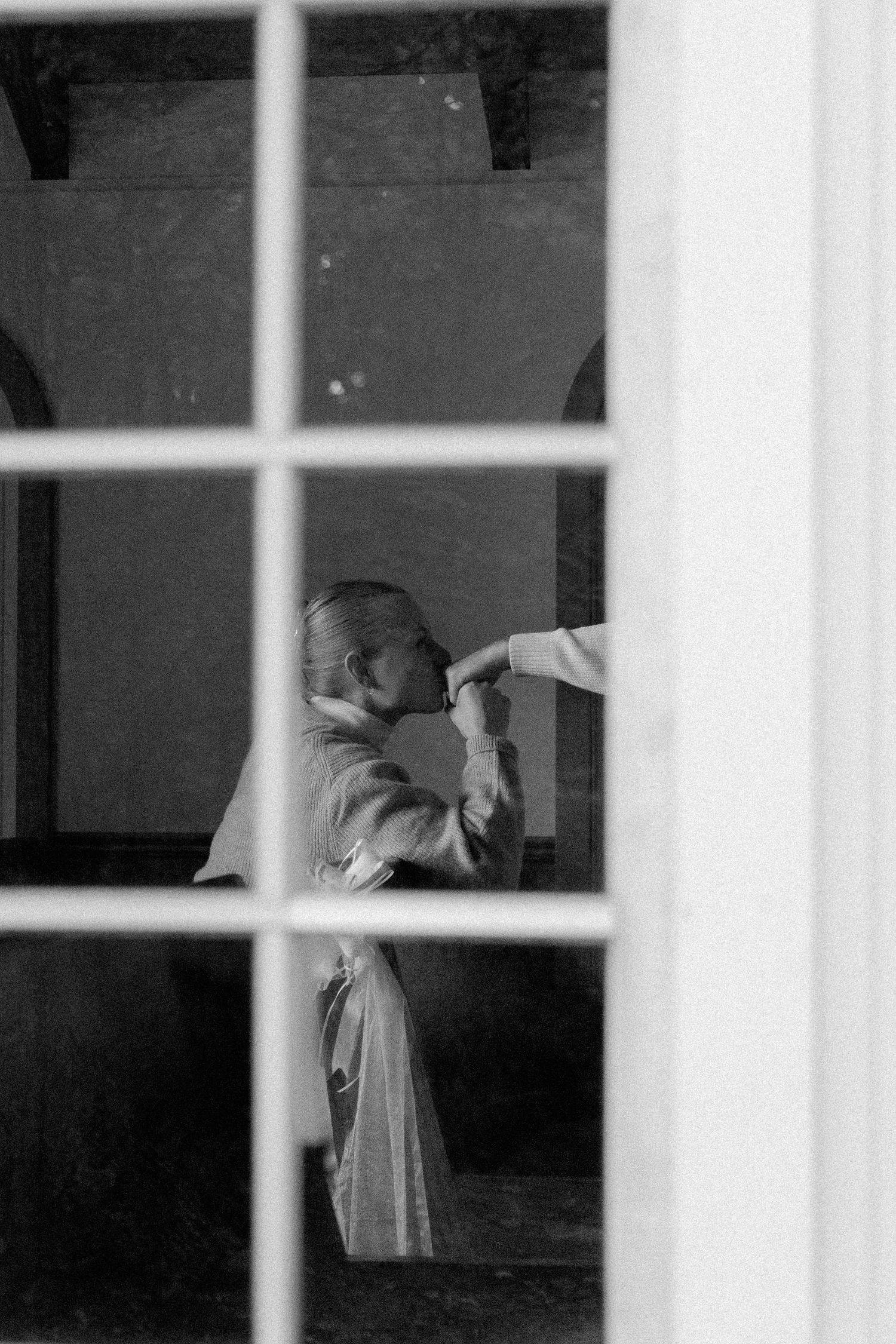 Black and white photo of child kissing someone's hand viewed through a window with white frames.