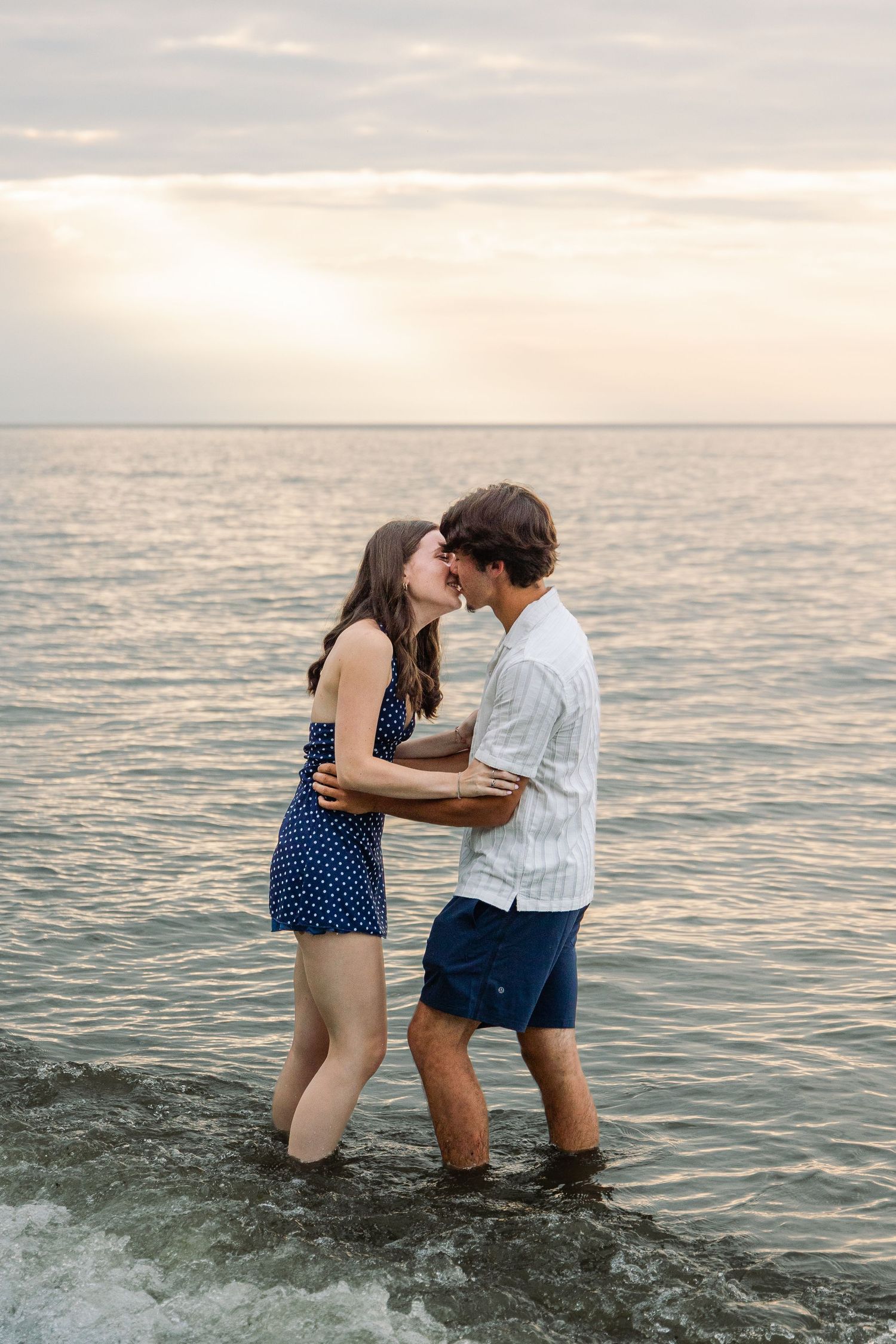 A couple shares a romantic kiss while standing in the ocean at sunset, wearing casual summer clothing.