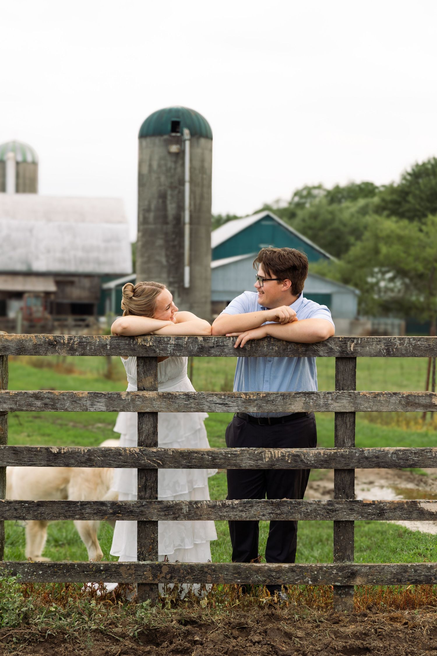 Couple leaning on rustic wooden fence gazing at each other with barn and silo in background.