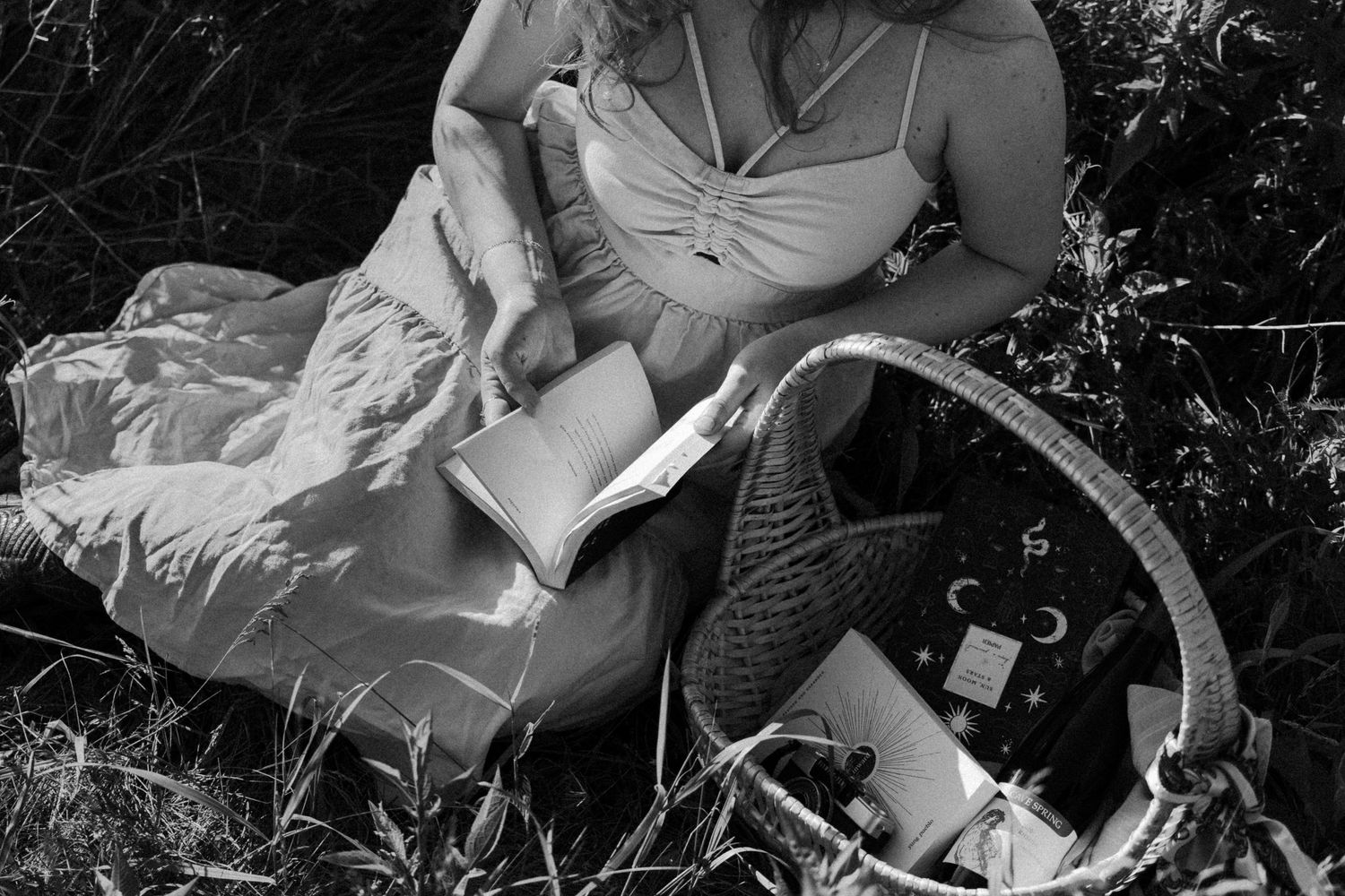 Black and white photo of a woman in a dress reading a book at an outdoor picnic.