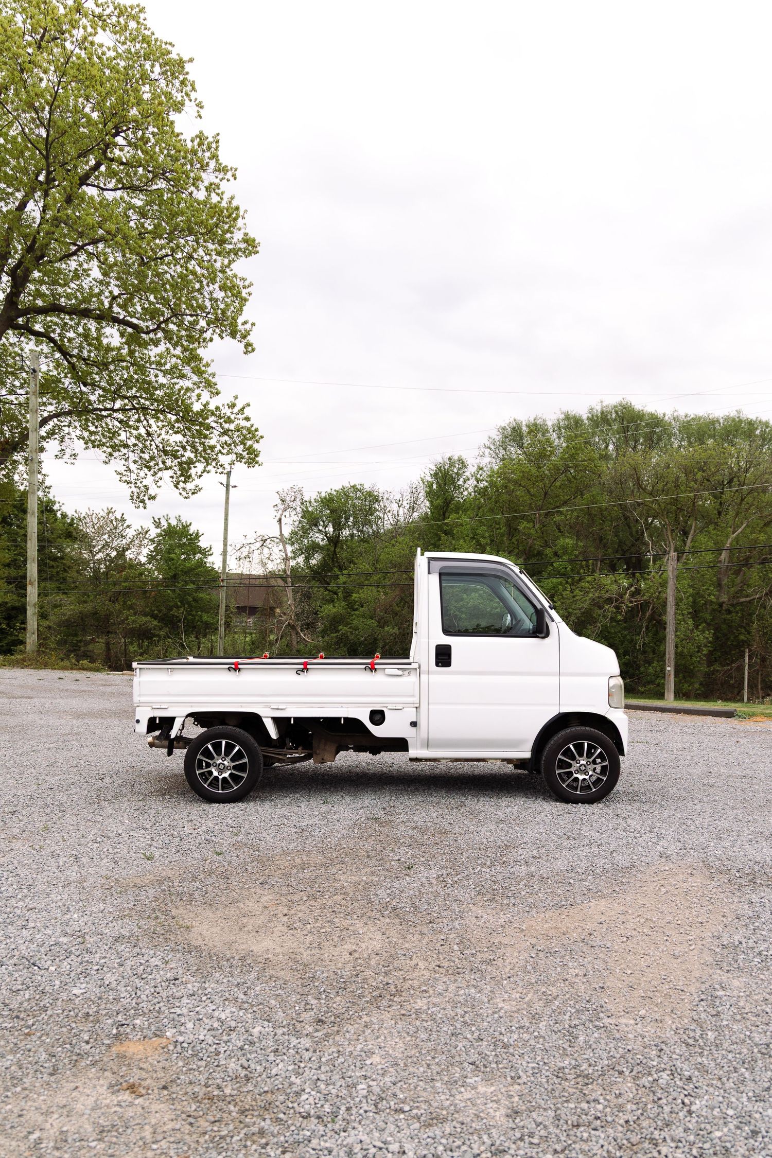 Small white Japanese mini truck parked on a gravel lot surrounded by green trees.