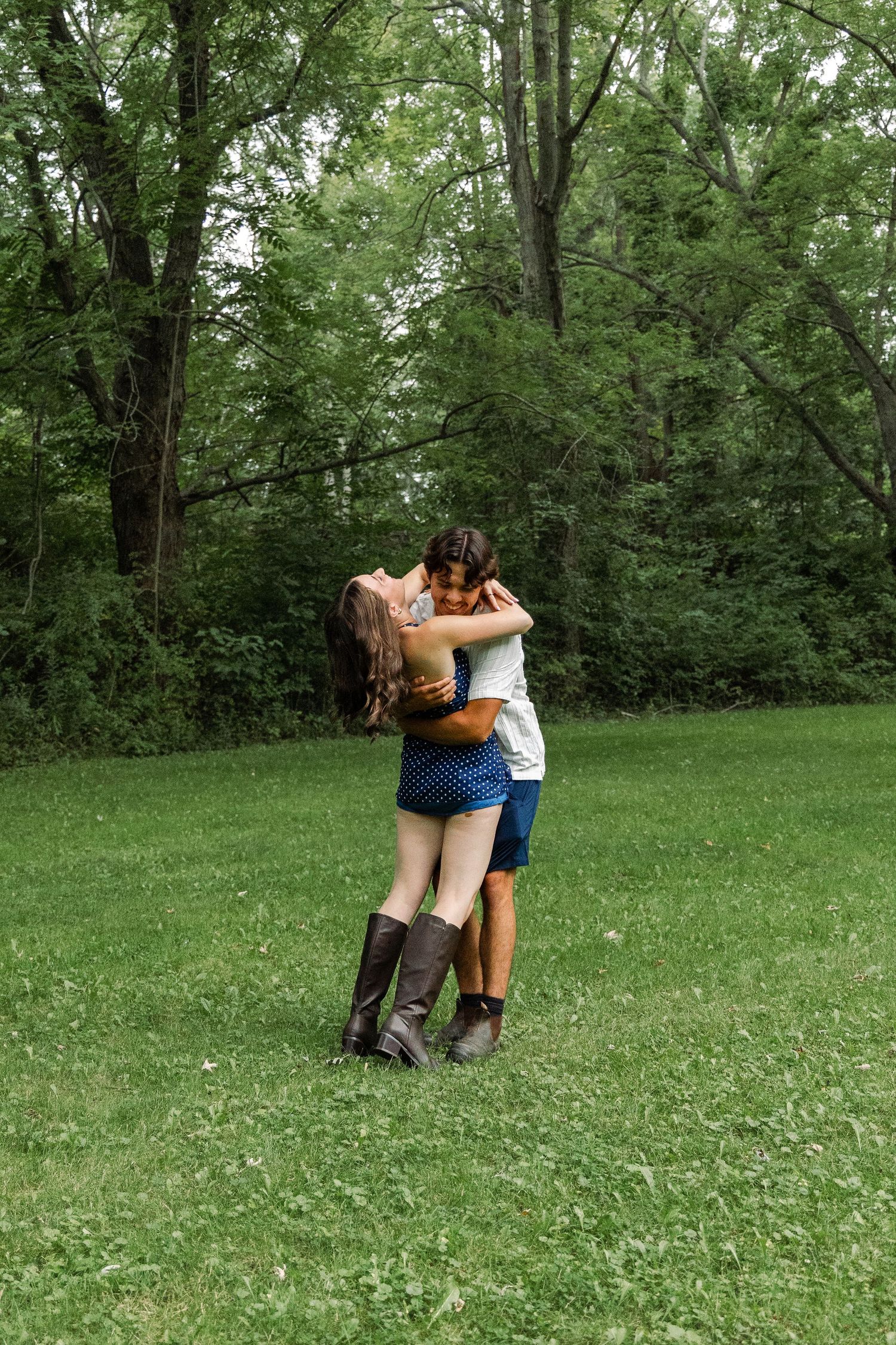 A couple embraces and kisses while standing on a green grassy lawn surrounded by lush trees.