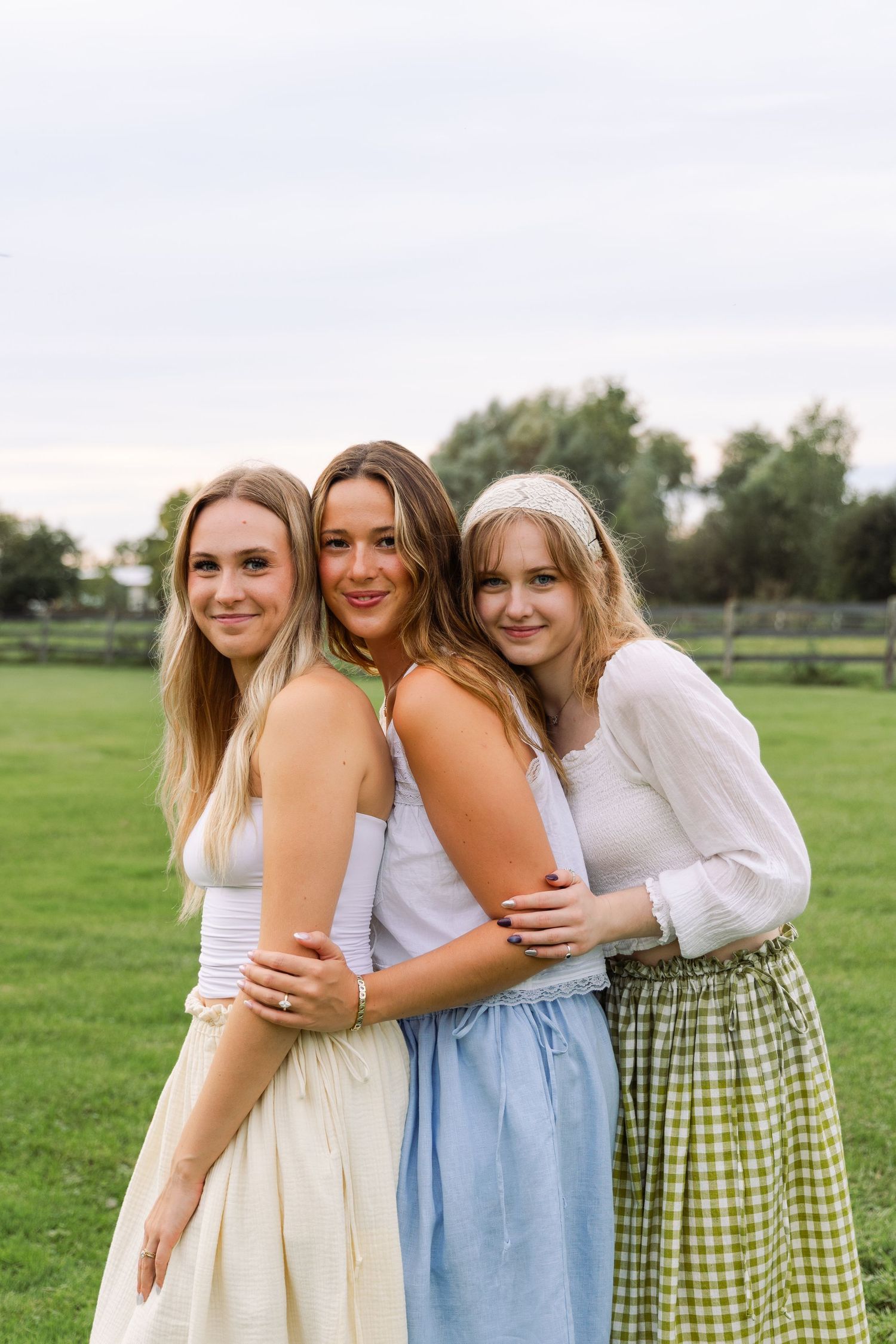 Three young women in flowy skirts embrace outdoors in a green field for a summer portrait.