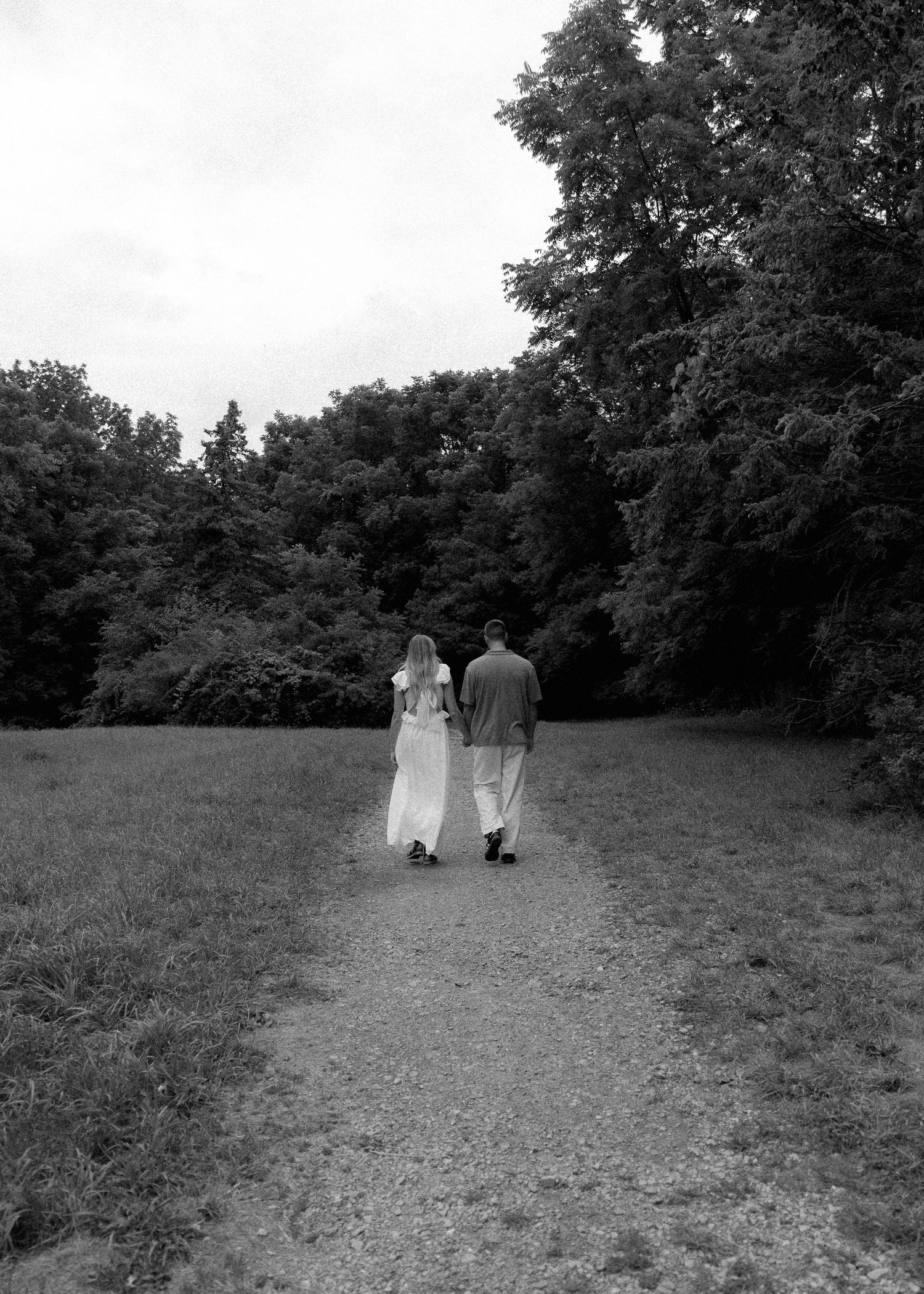 Black and white photo of couple walking hand in hand down tree-lined path in serene park.