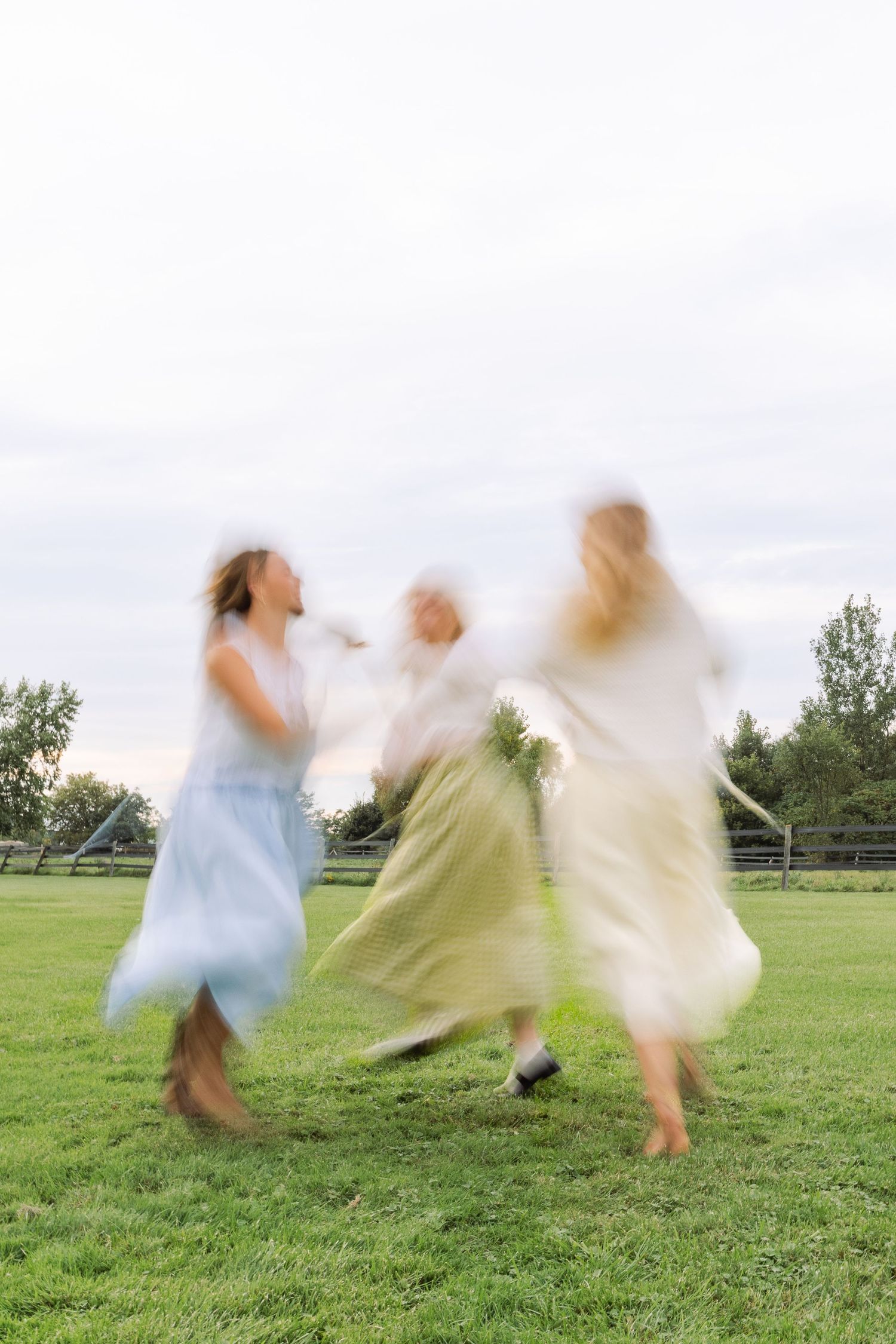Motion-blurred photo of three women in pastel midi skirts joyfully spinning together on a green lawn.