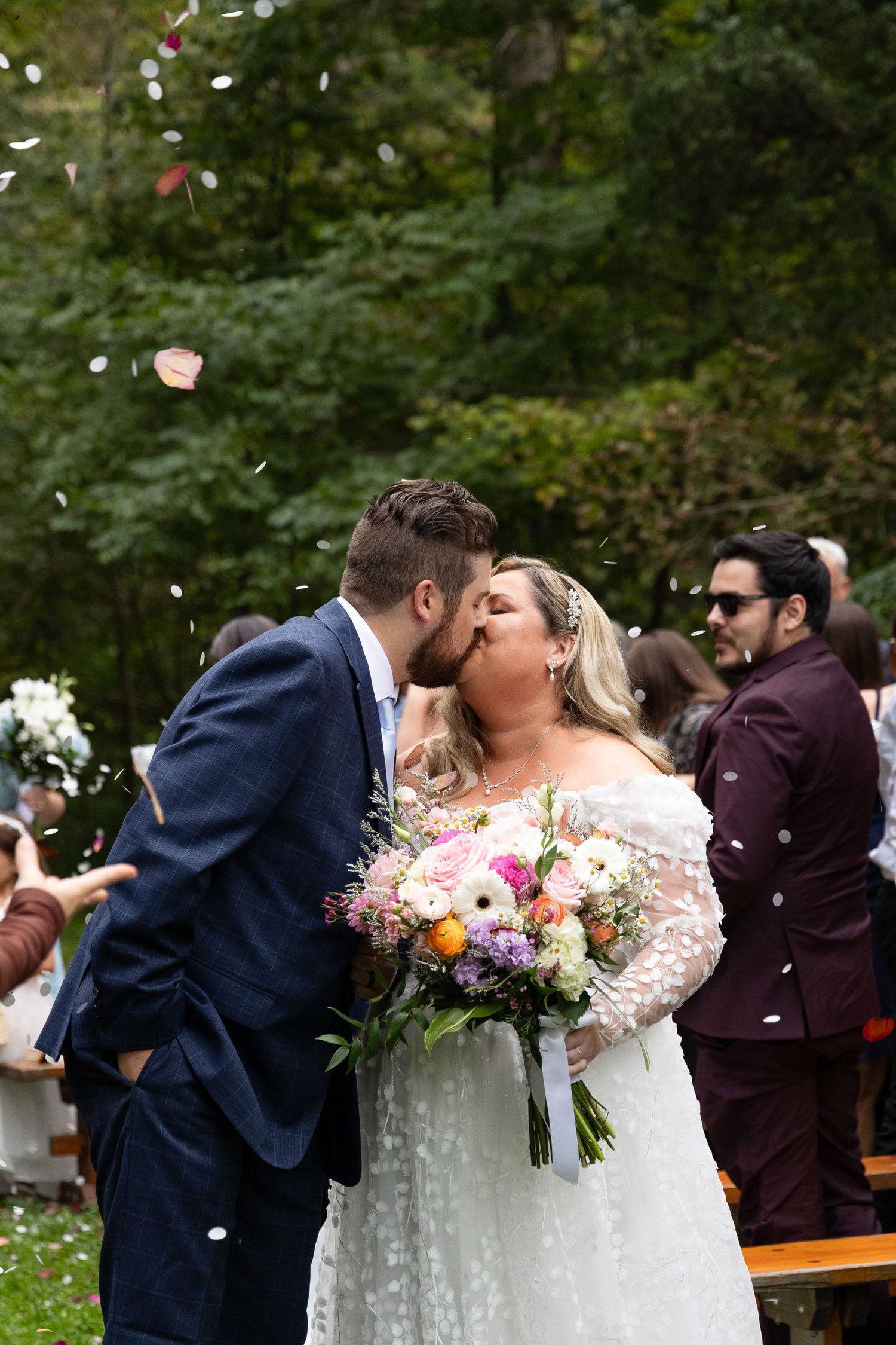Bride and groom share first kiss as confetti falls during outdoor woodland wedding ceremony.