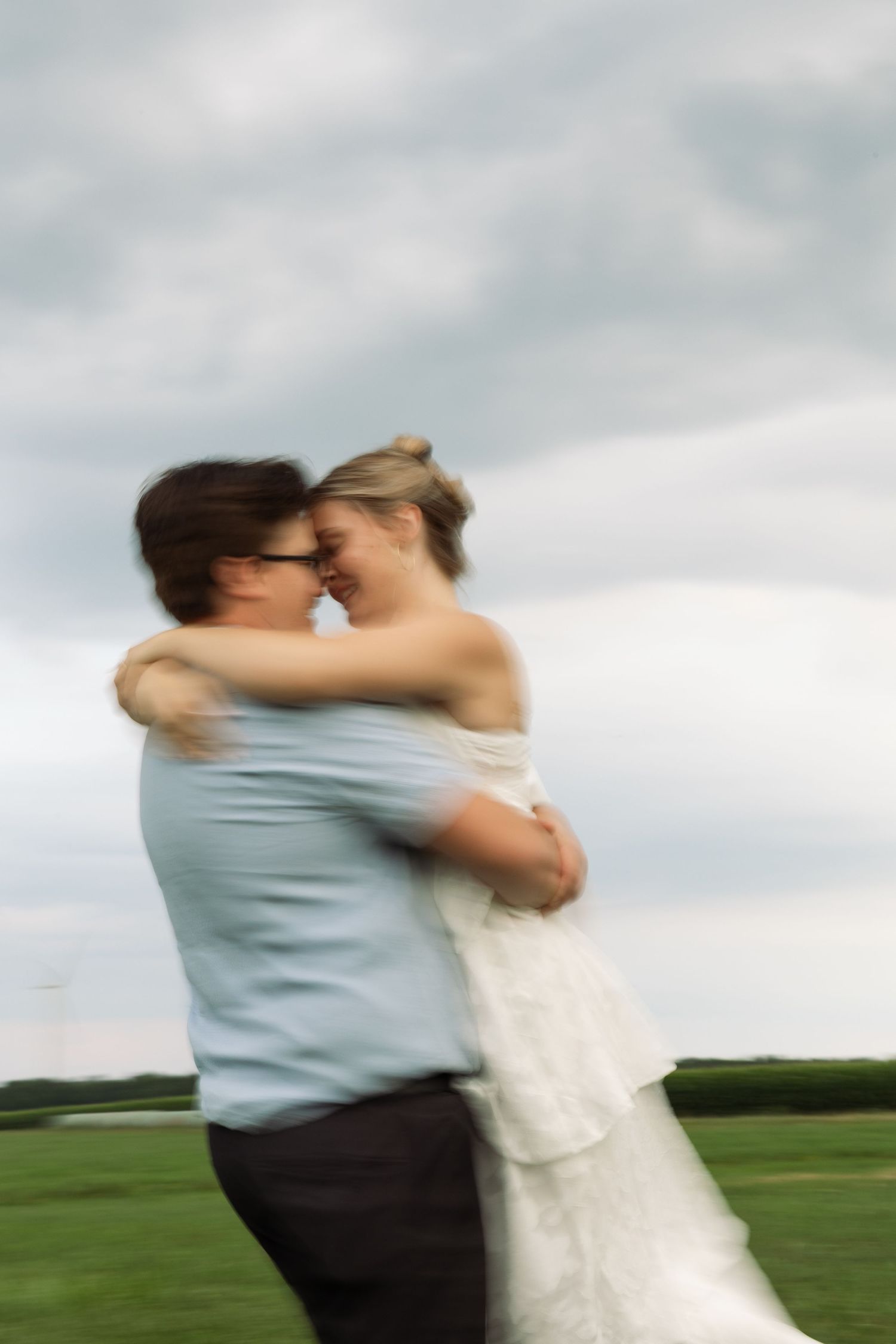 Blurred motion photo of a couple embracing and touching foreheads in a green field under a cloudy sky.