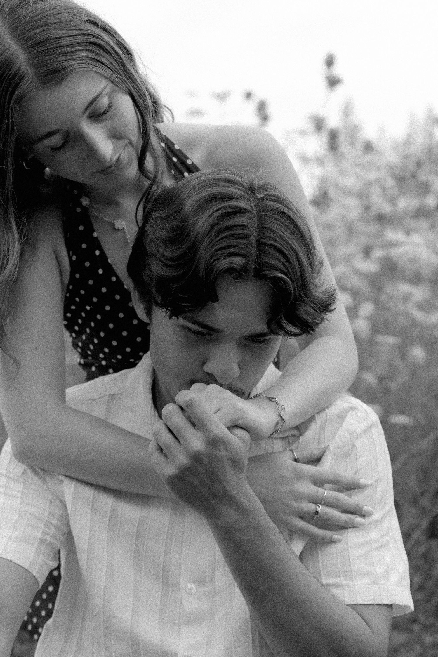 Black and white photo of a young man kissing a woman's hand while she embraces him from behind.