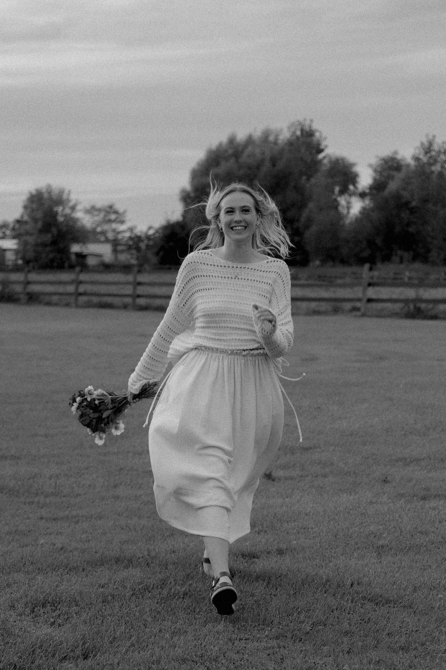 Black and white photo of a smiling blonde woman running through a field holding a bouquet of wildflowers.