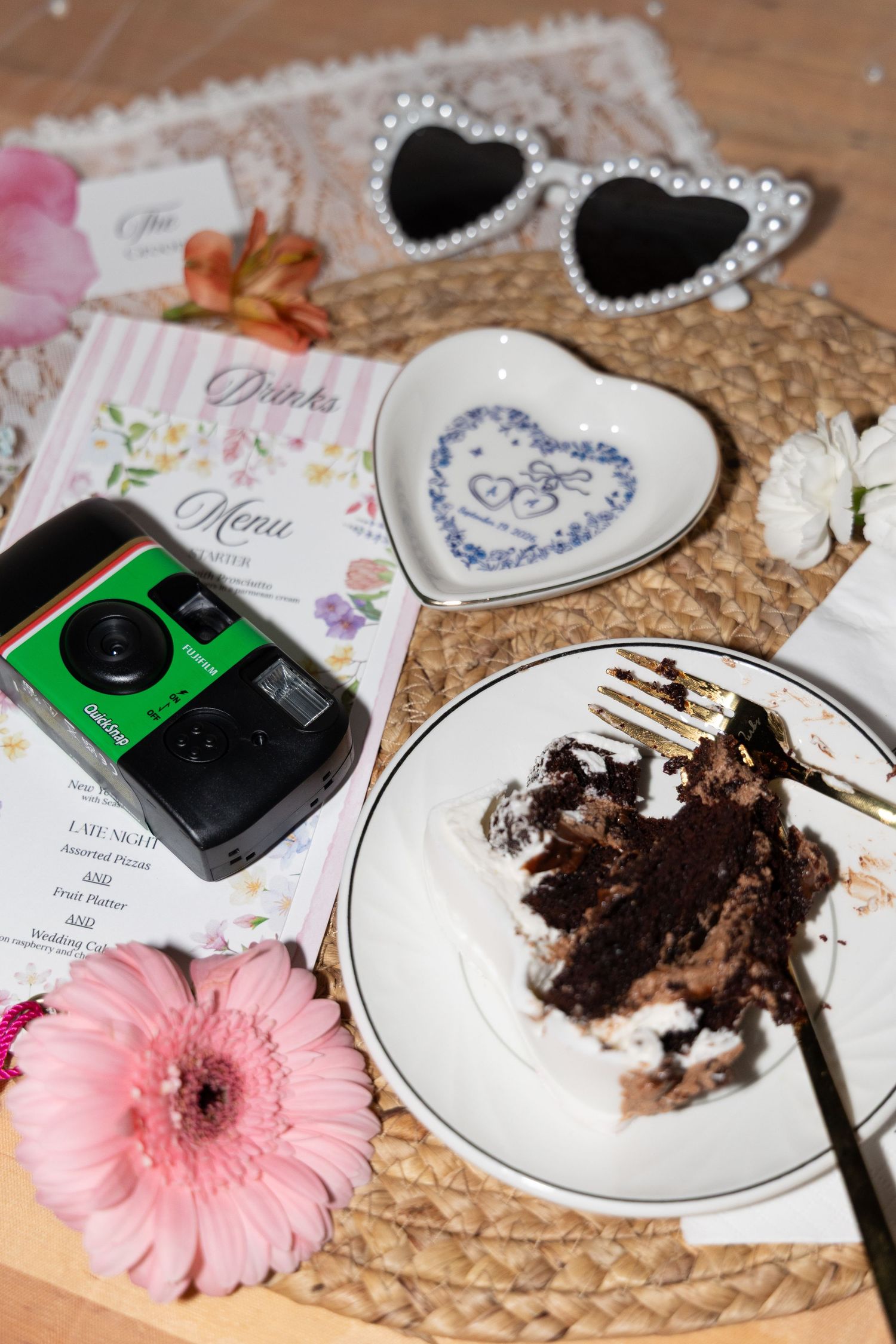 Flat lay of wedding details including chocolate cake, disposable camera, drinks menu, heart dish, and pearl sunglasses on wicker mat.