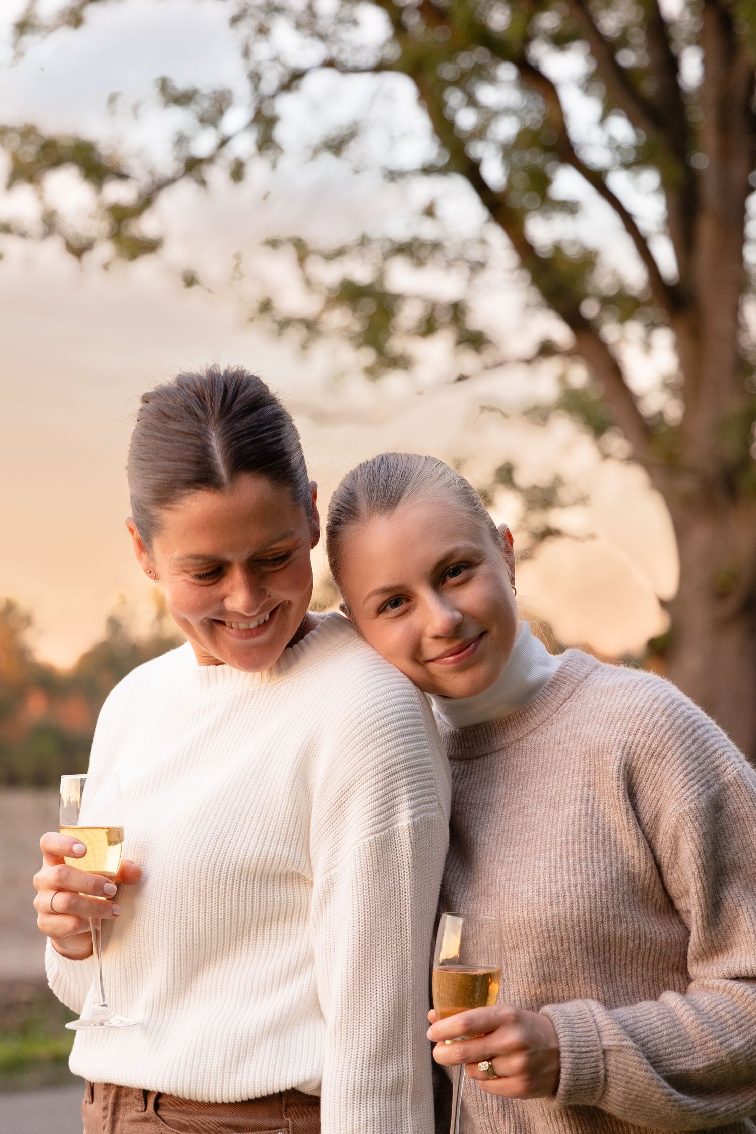 Two smiling women in matching cream sweaters holding champagne glasses at sunset outdoors.