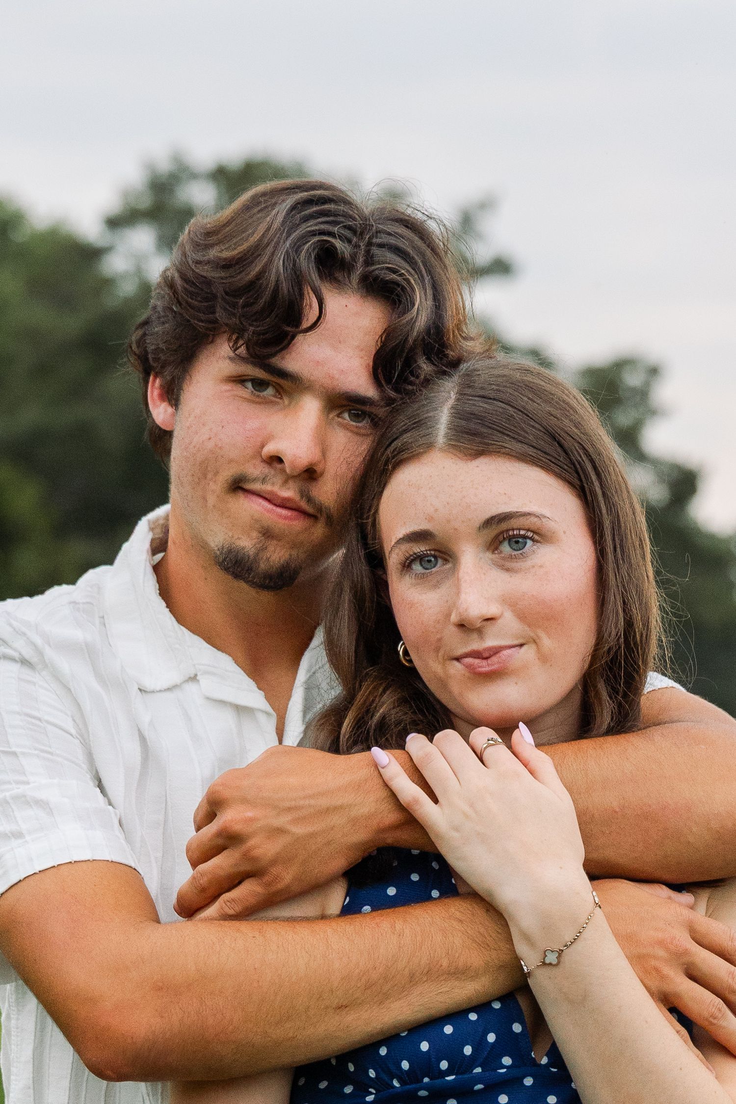A young couple poses together outdoors, the man embracing the woman from behind near green trees.