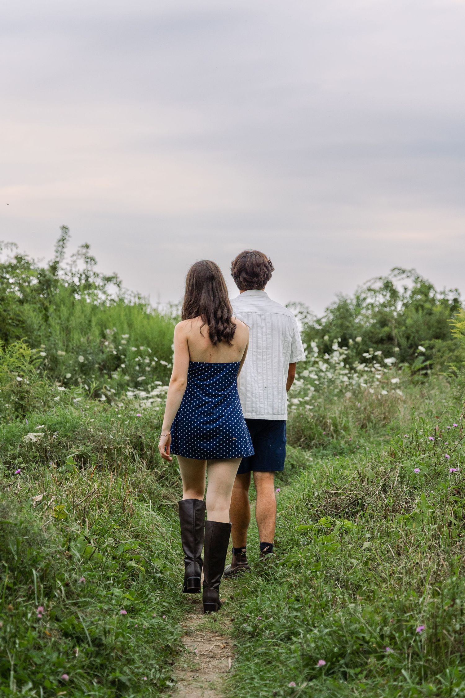 A couple walks hand in hand down a narrow path through a wildflower meadow under a cloudy sky.