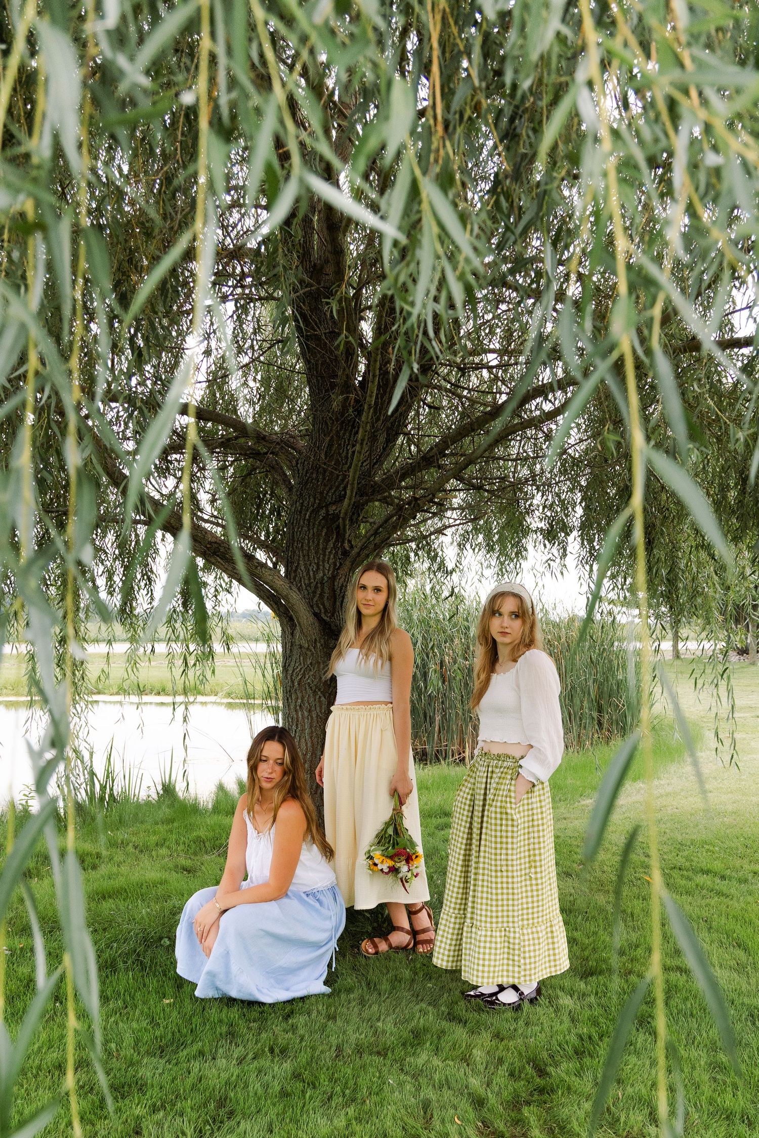 Three women in maxi skirts pose under a weeping willow tree near a serene lake.