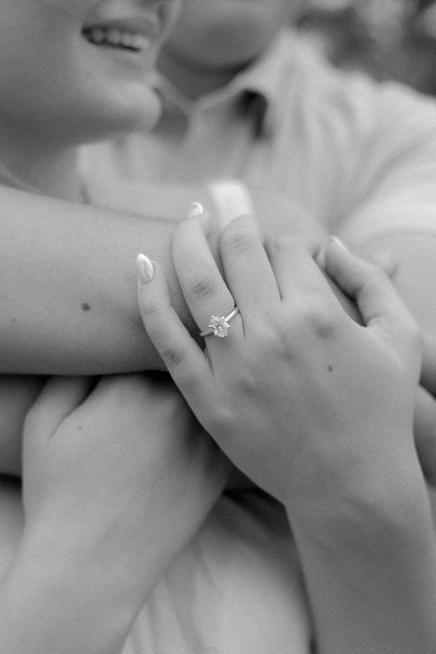 Black and white close-up of woman's hand displaying a solitaire diamond engagement ring while being embraced.