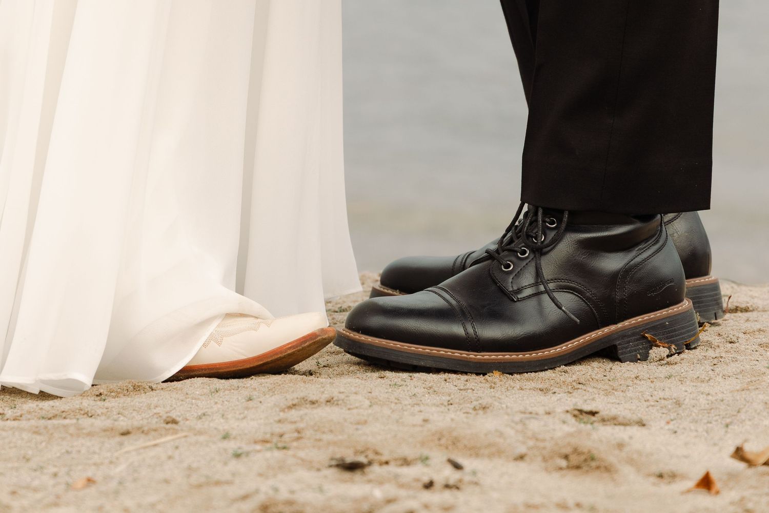 Close-up of groom wearing black leather oxford shoes standing beside bride in white dress on sandy ground.