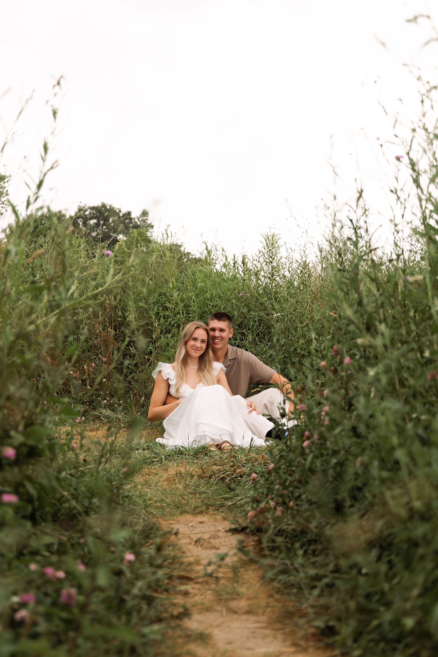 Happy couple sitting together on a dirt path surrounded by tall green wildflowers.