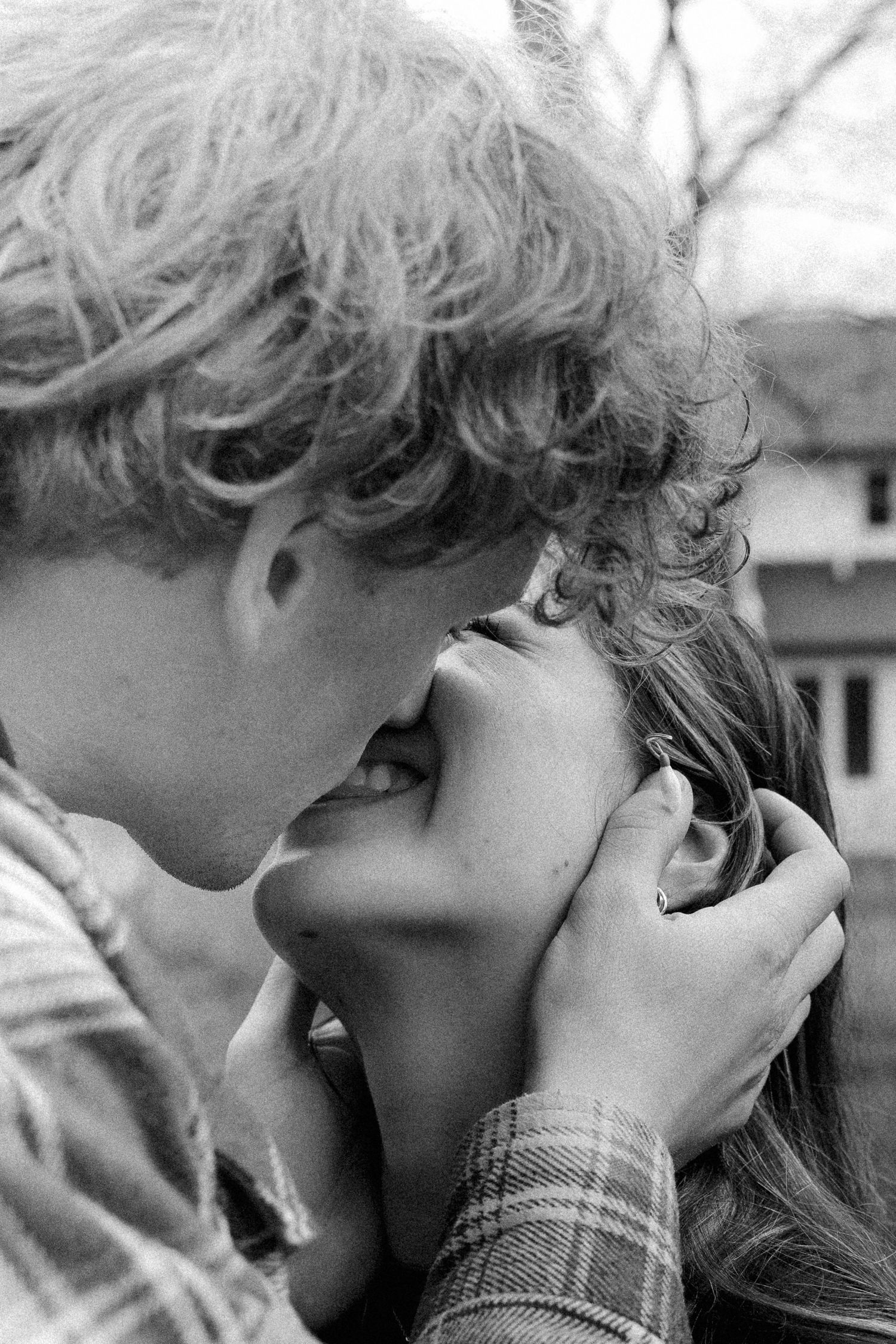 Black and white photo of a young couple sharing a tender kiss outdoors in a plaid shirt.