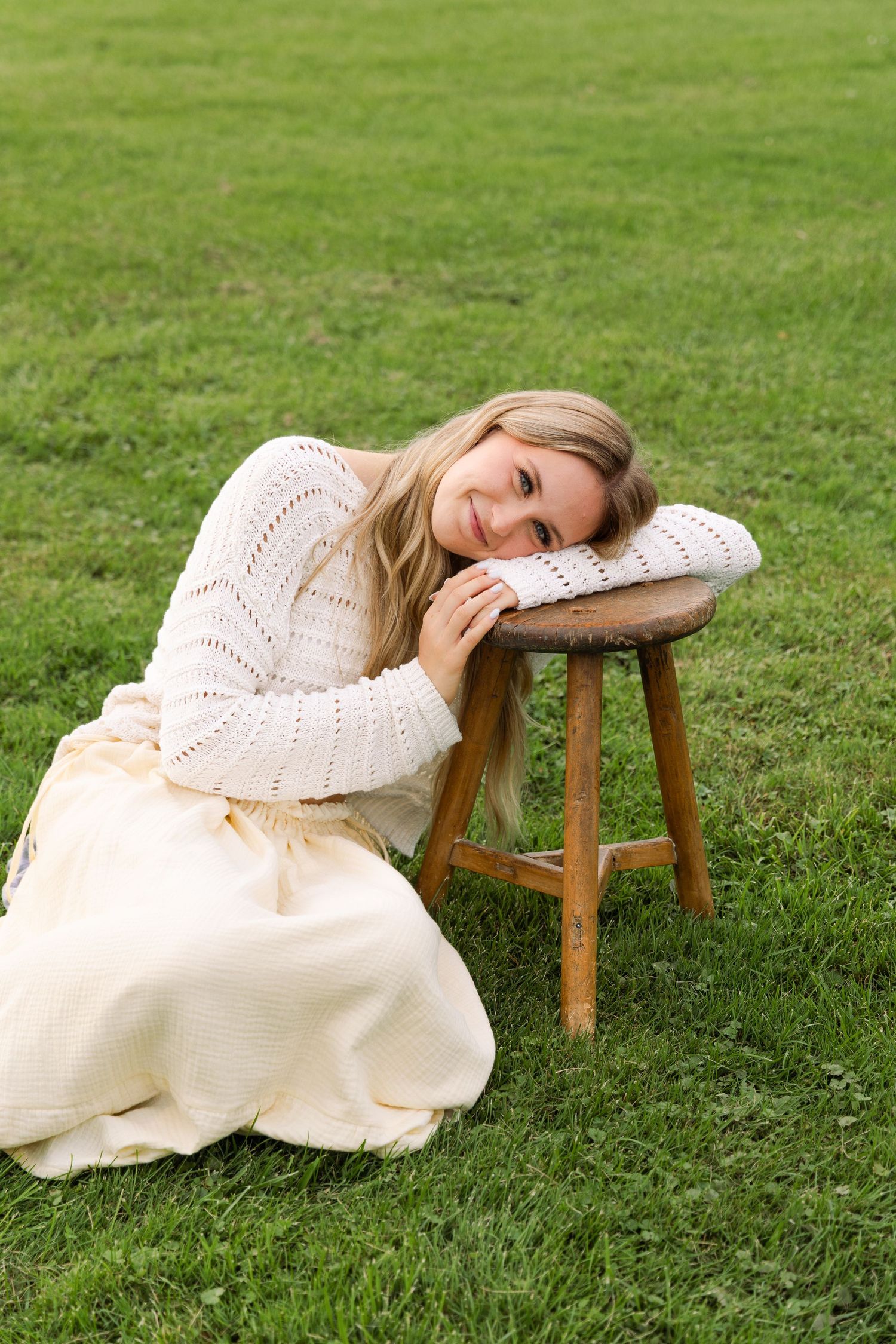 A smiling blonde woman in a cream knit sweater and ivory midi skirt rests her head on a wooden stool outdoors.