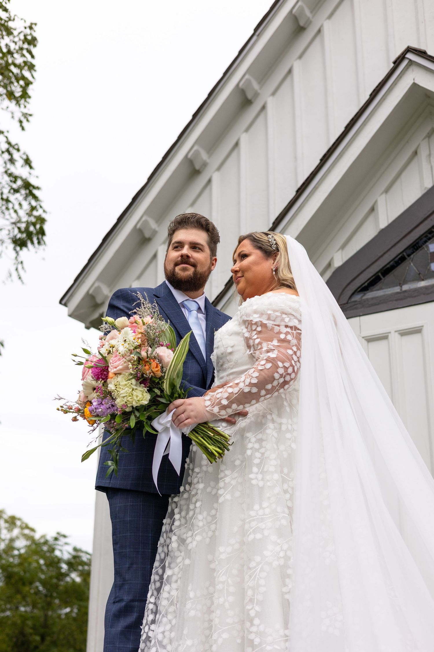 Bride and groom pose with colorful floral bouquet outside white barn wedding venue.