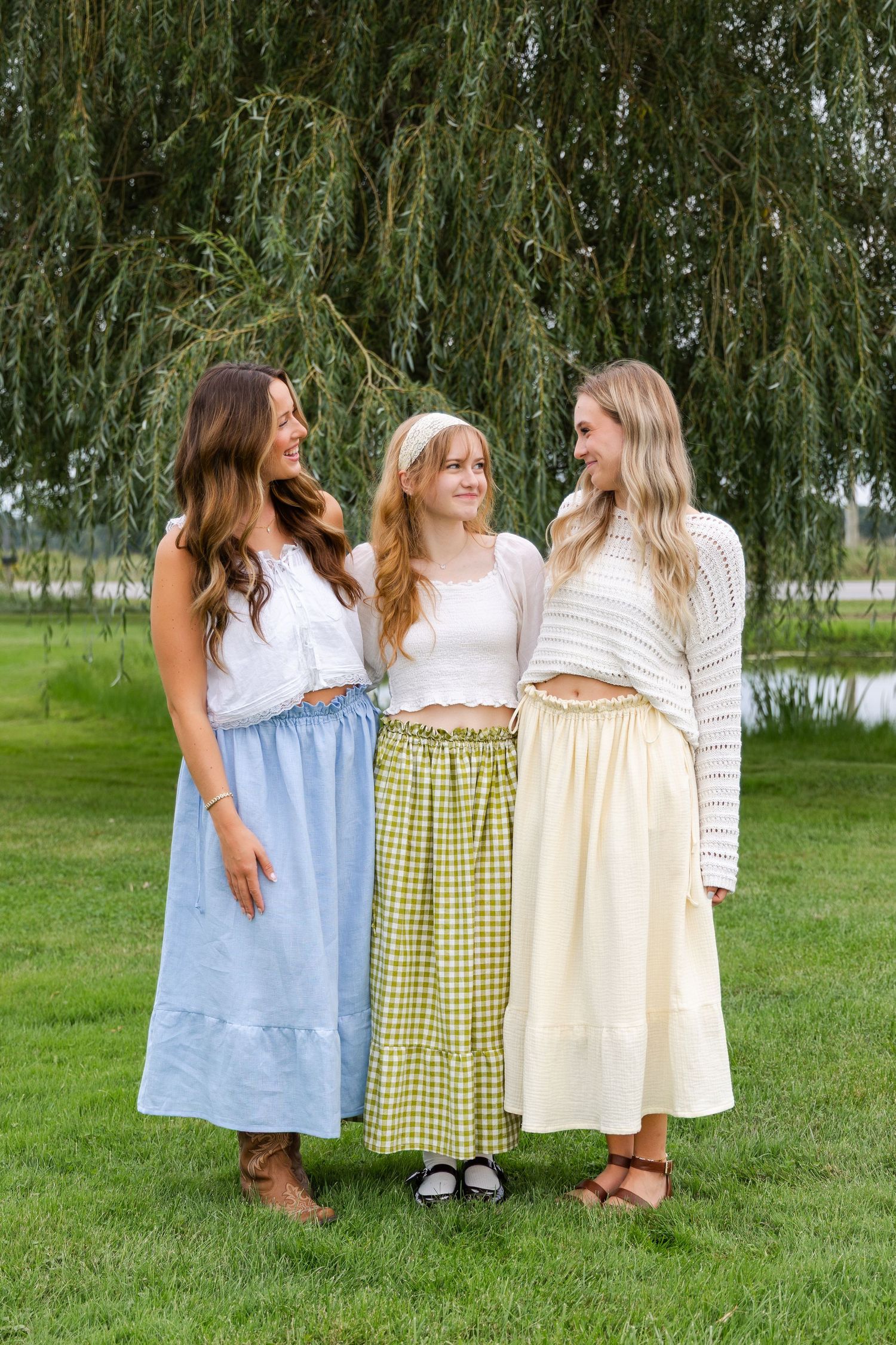 Three young women in white tops and pastel midi skirts smile together outdoors near a weeping willow tree.
