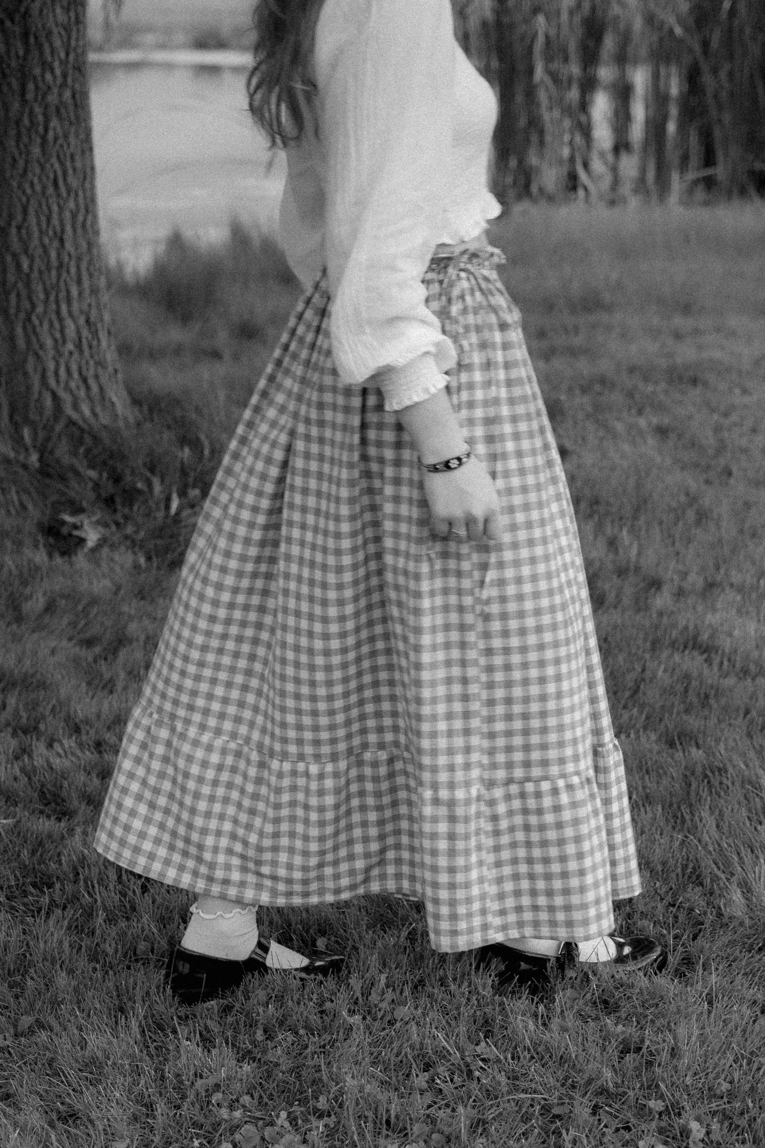 Black and white photo of a girl wearing a gingham midi skirt and white blouse standing in a grassy field.