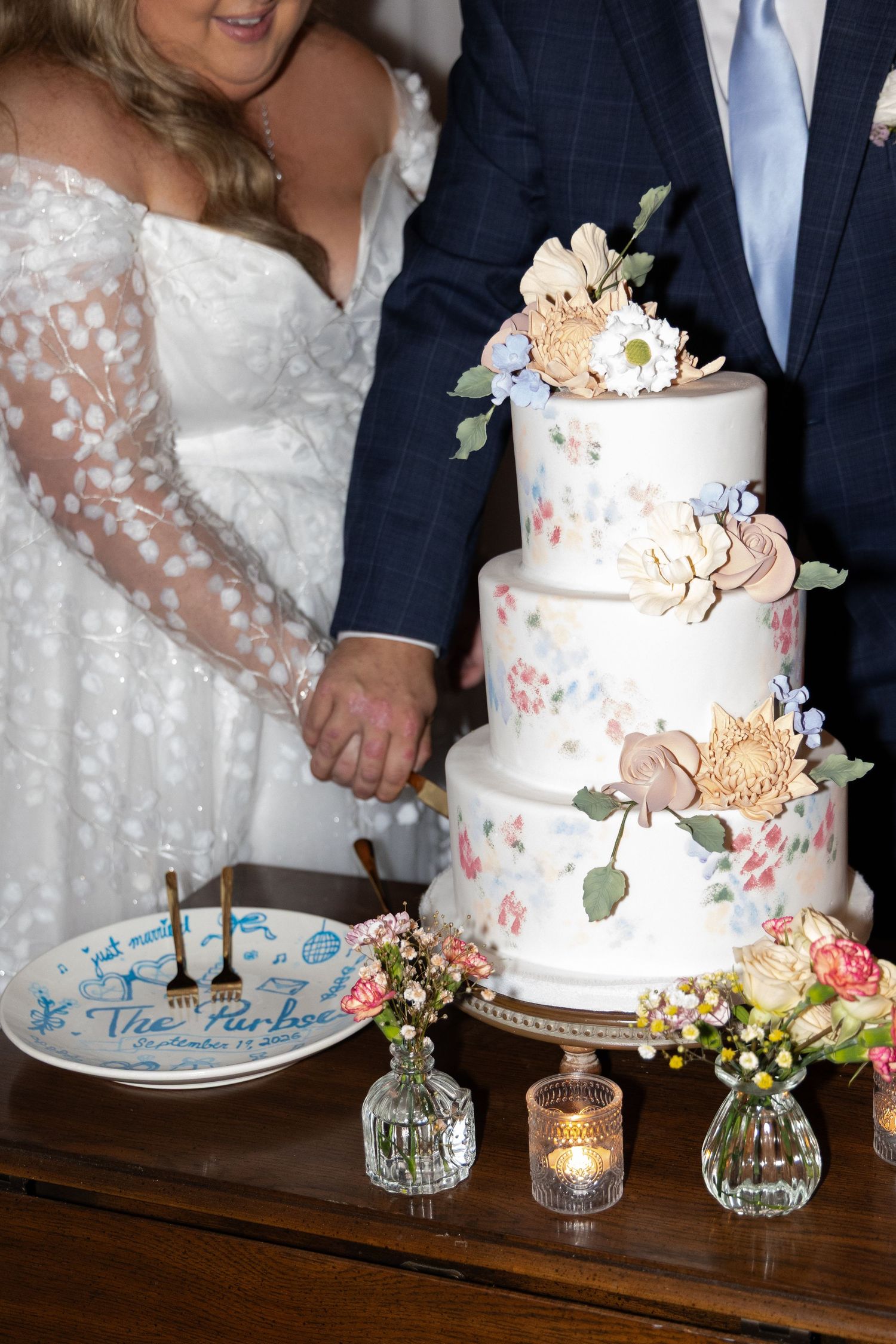 Bride and groom cut three-tier floral wedding cake decorated with painted roses and flowers.
