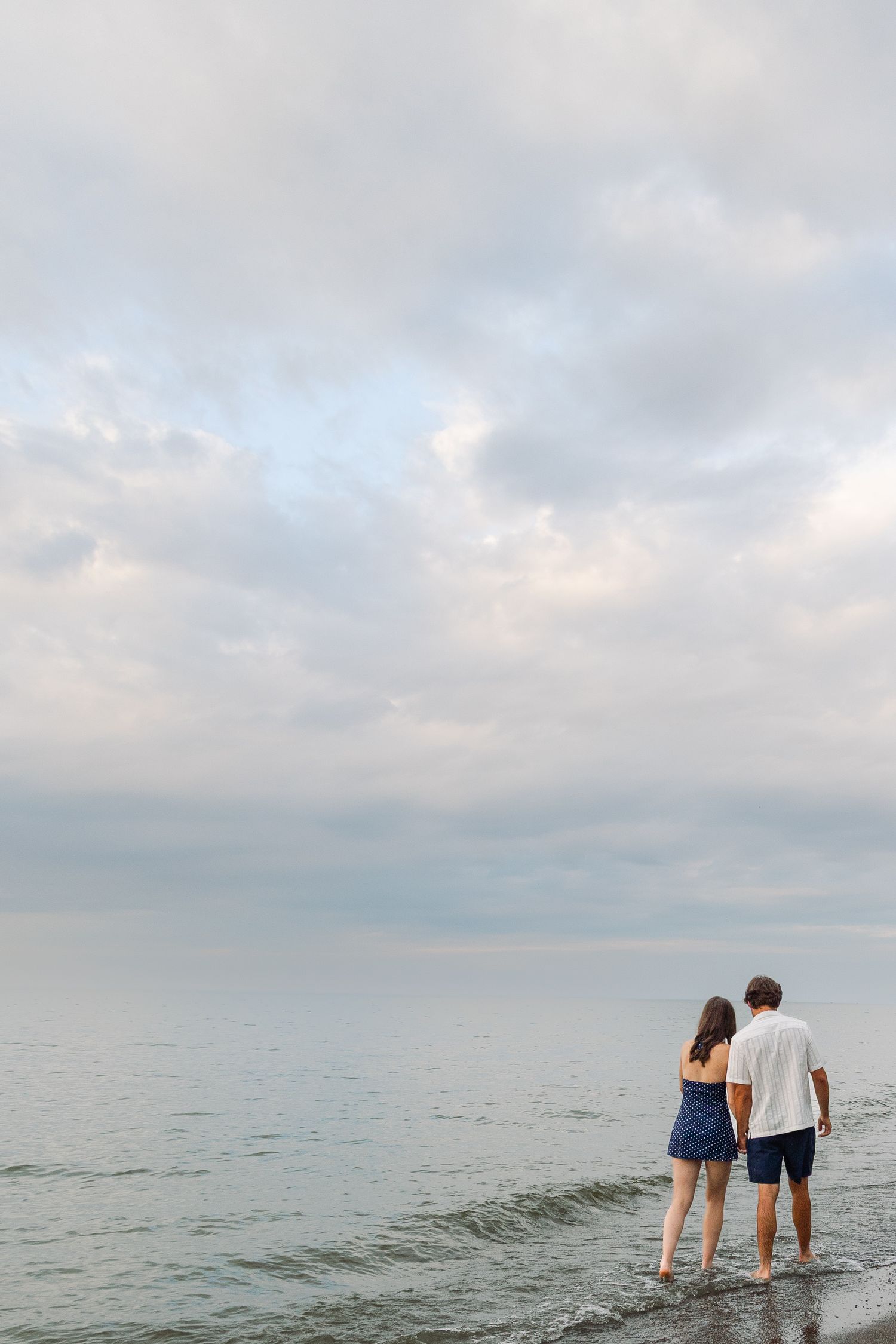 A couple stands barefoot at the water's edge on a beach under a dramatic cloudy sky at dusk.