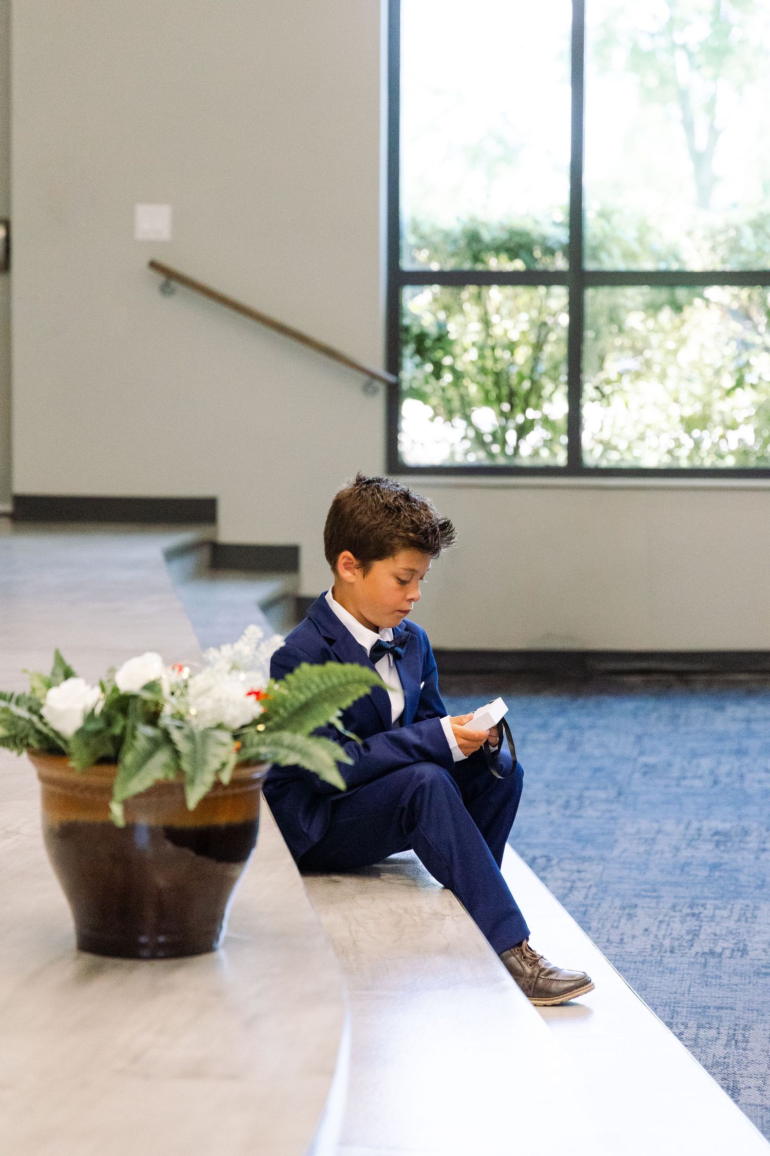 A young boy in a navy suit sits on a bench looking at a card next to a floral arrangement indoors.