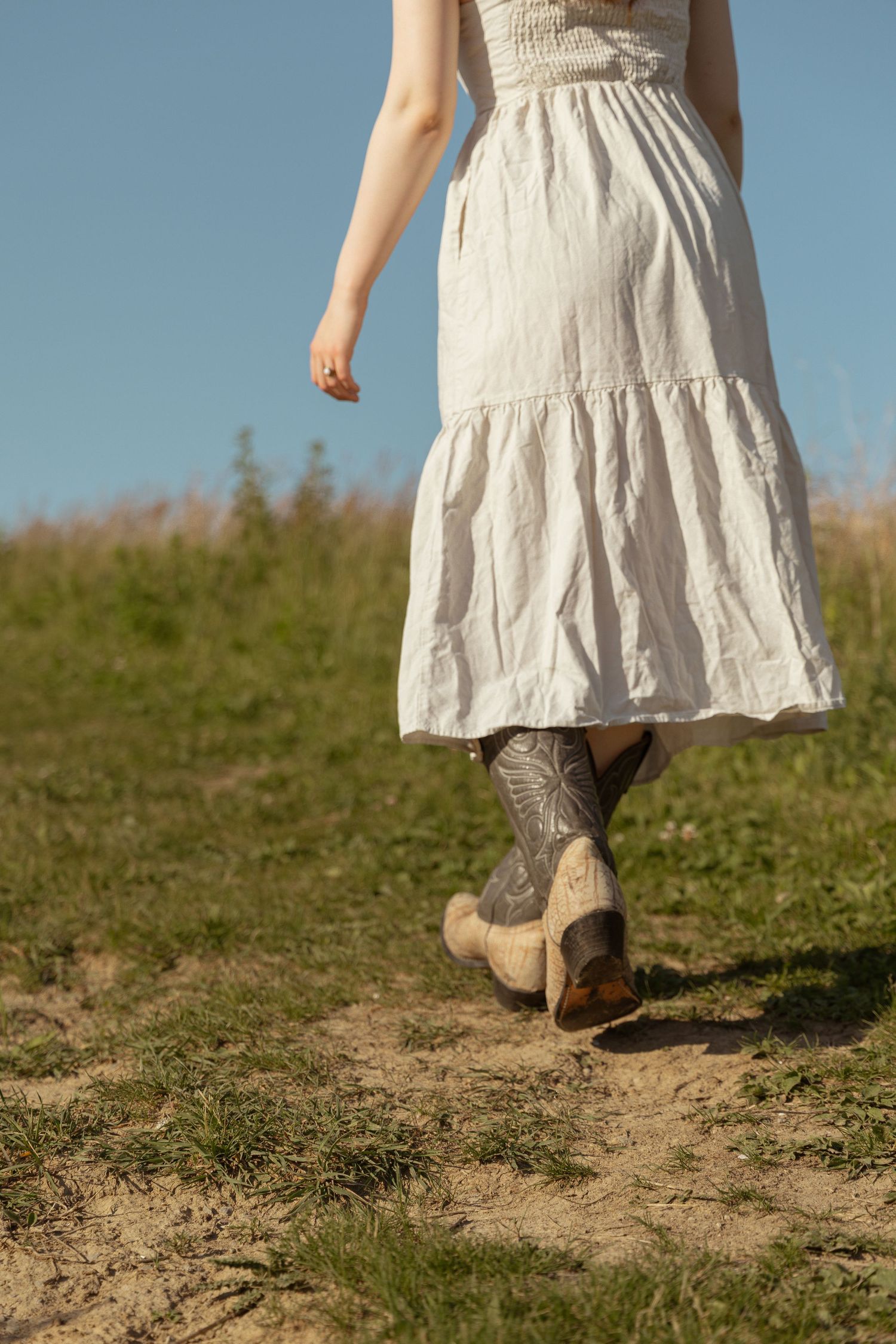 Woman in white linen dress and cowboy boots walking on rural dirt path in sunny field.