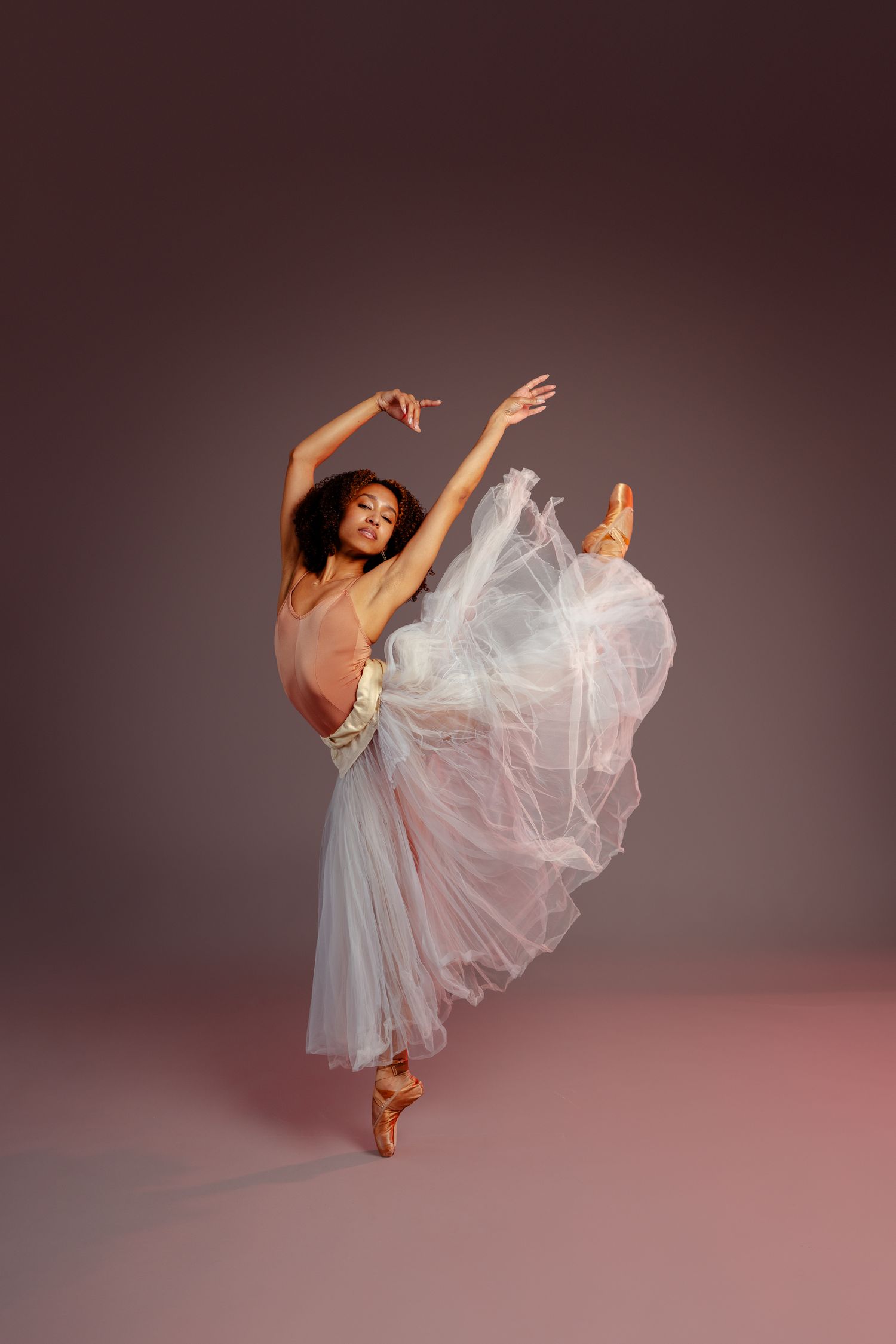Graceful ballerina in white tutu dress dancing en pointe against a mauve studio background.