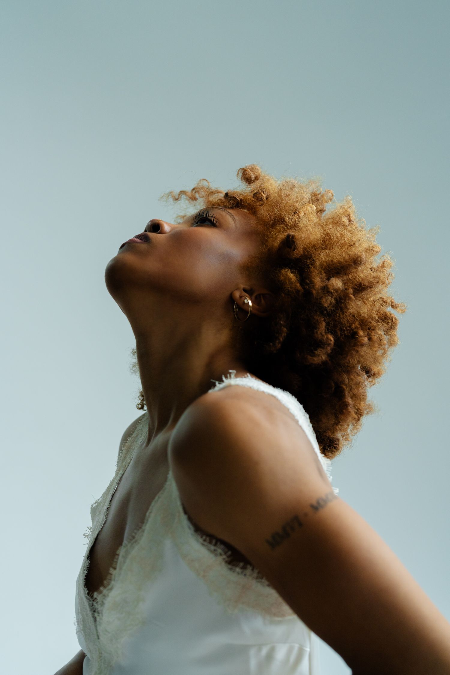 A Black woman with curly blonde hair tilts her head back wearing a white sleeveless top against a light blue background.