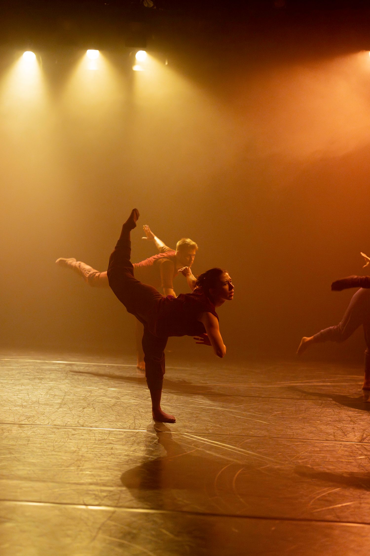 Dancer performs dramatic high kick on dimly lit stage with warm amber spotlights creating atmospheric silhouette effect.