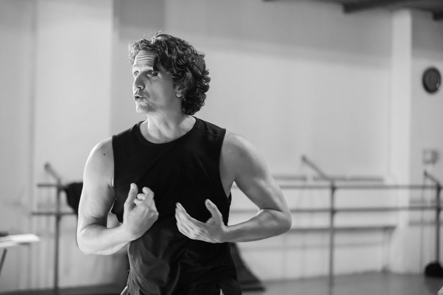 A dancer in a black tank top practices expressive movements in a bright dance studio.